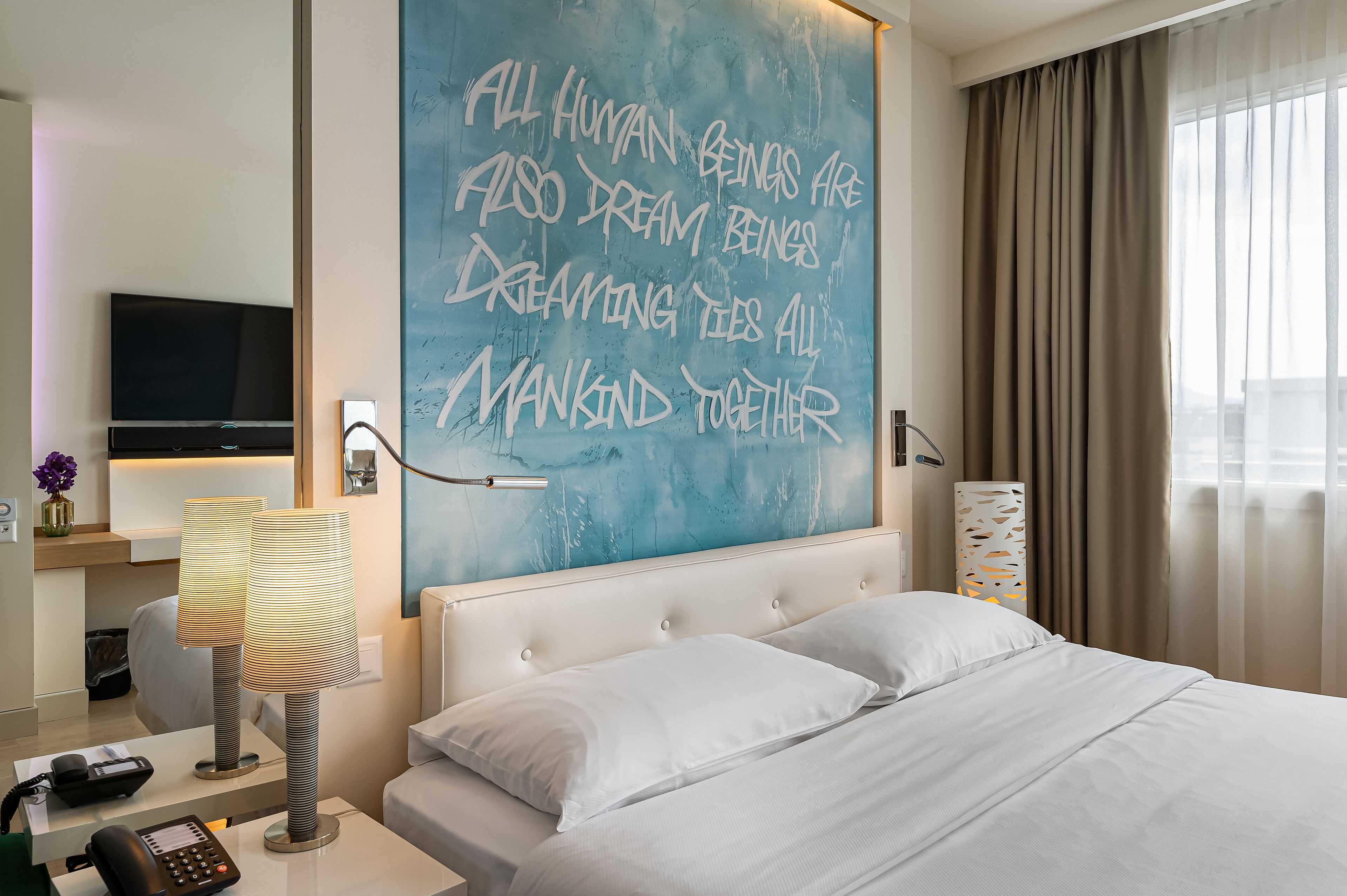 Habitación de hotel con una cama grande, un escritorio con teléfono, una lámpara, un televisor de pantalla plana y una cita motivacional en la pared.