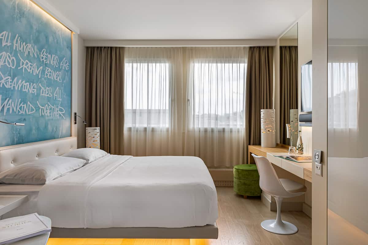 A modern hotel room with a large bed, a desk, and a chair. There is a large window with curtains, a mirror, and a wall with a motivational quote.