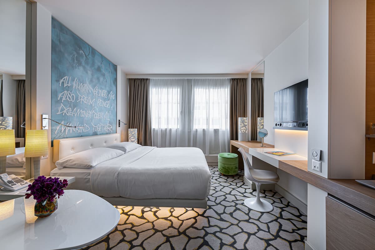 A modern hotel room with a large bed, a desk with a chair, a round table with a vase of flowers, a large window with curtains, and a blue wall with text.
