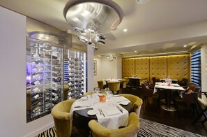 Un comedor con una estantería para vinos, mesas redondas y sillas tapizadas en amarillo.