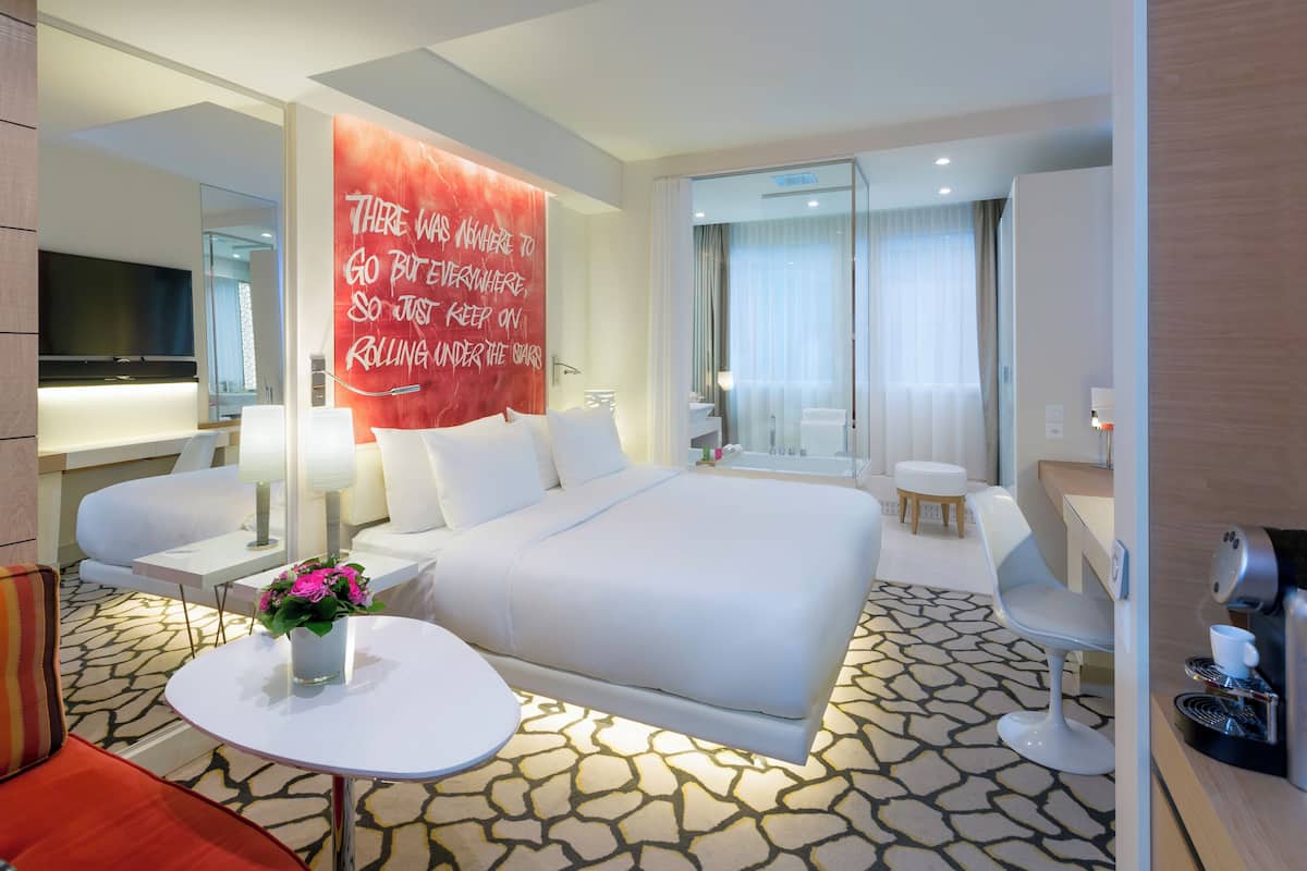 A modern hotel room with a large bed, a sitting area with a table and chairs, a bathroom with a shower and toilet, and a large mirror.
