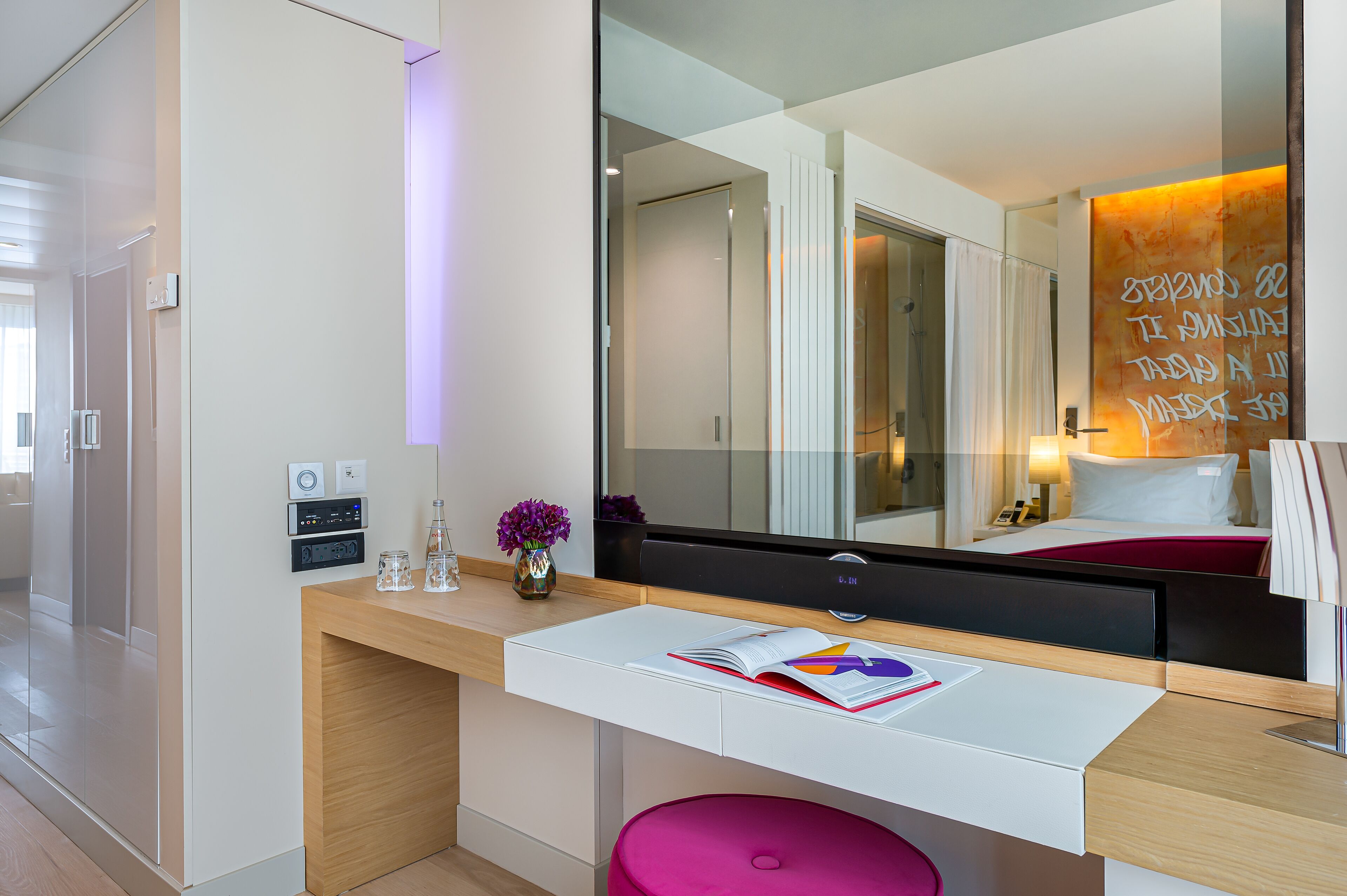 A modern hotel room with a white desk, a large mirror, and a pink stool.