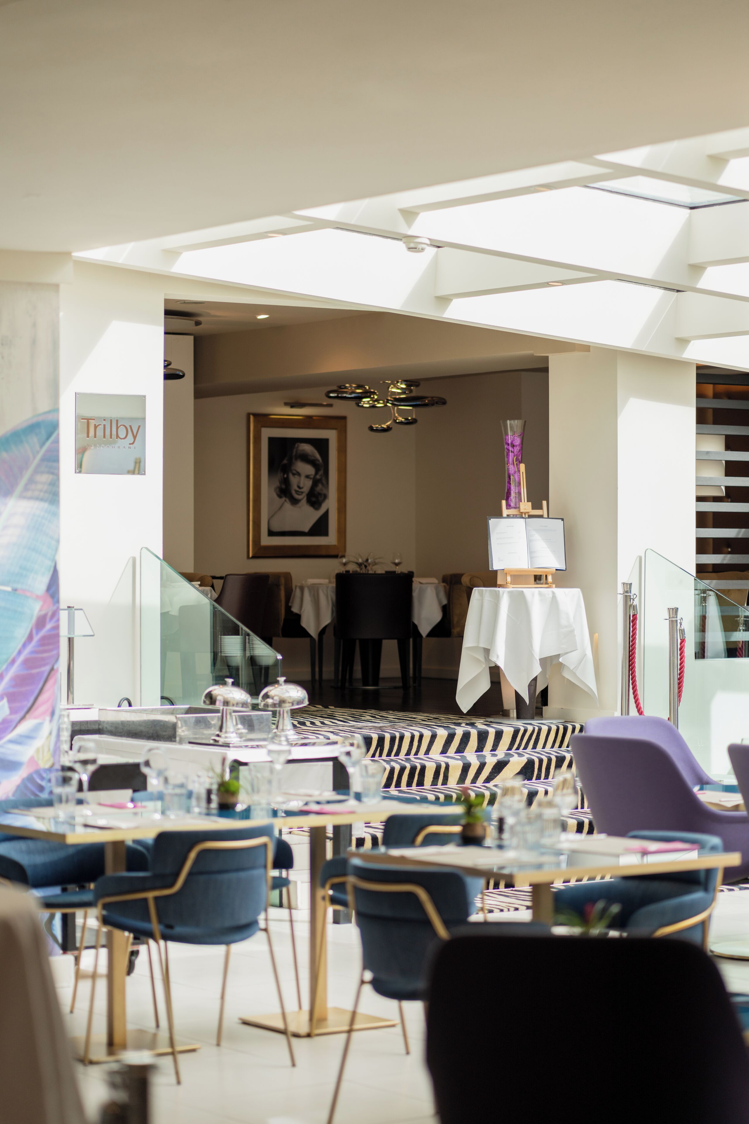 An indoor dining area with tables and chairs, a staircase leading to an upper level, and a framed portrait on the wall.