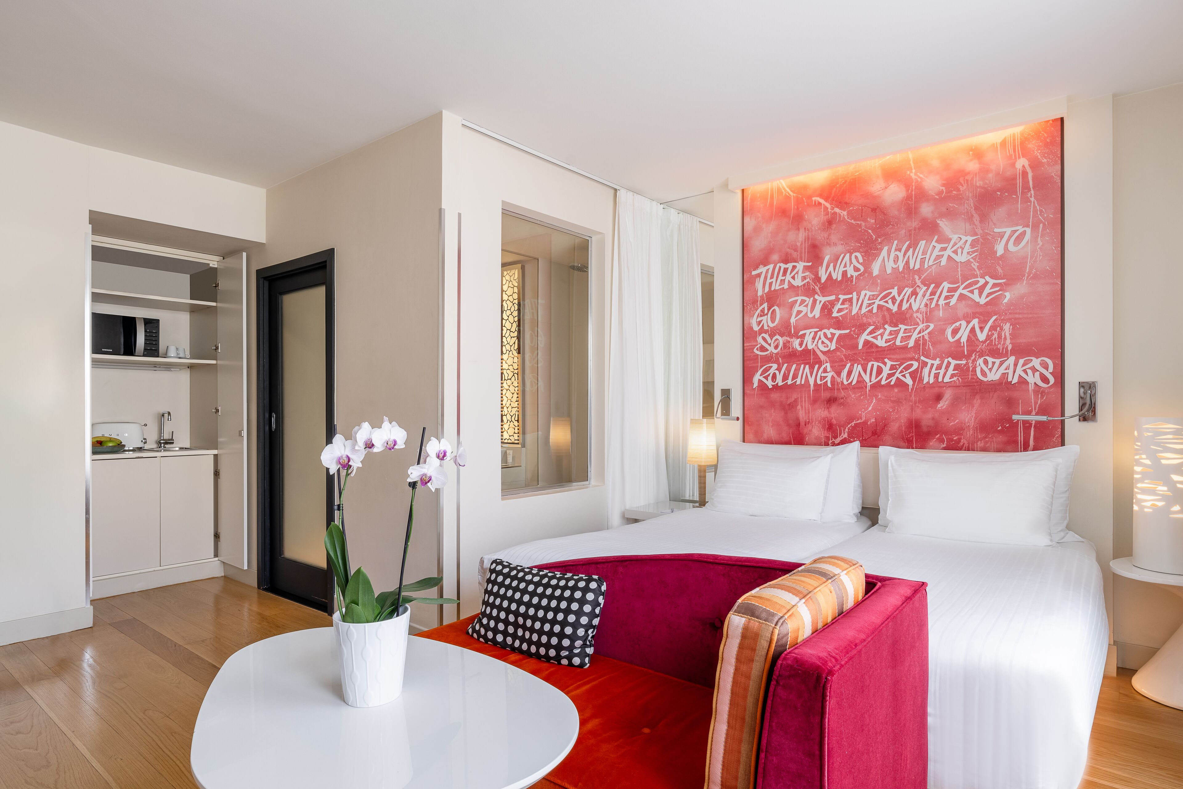 Een hotelkamer met een bed, een rode bank, een kleine tafel met een bloevas en een groot motiverend citaat op de muur.