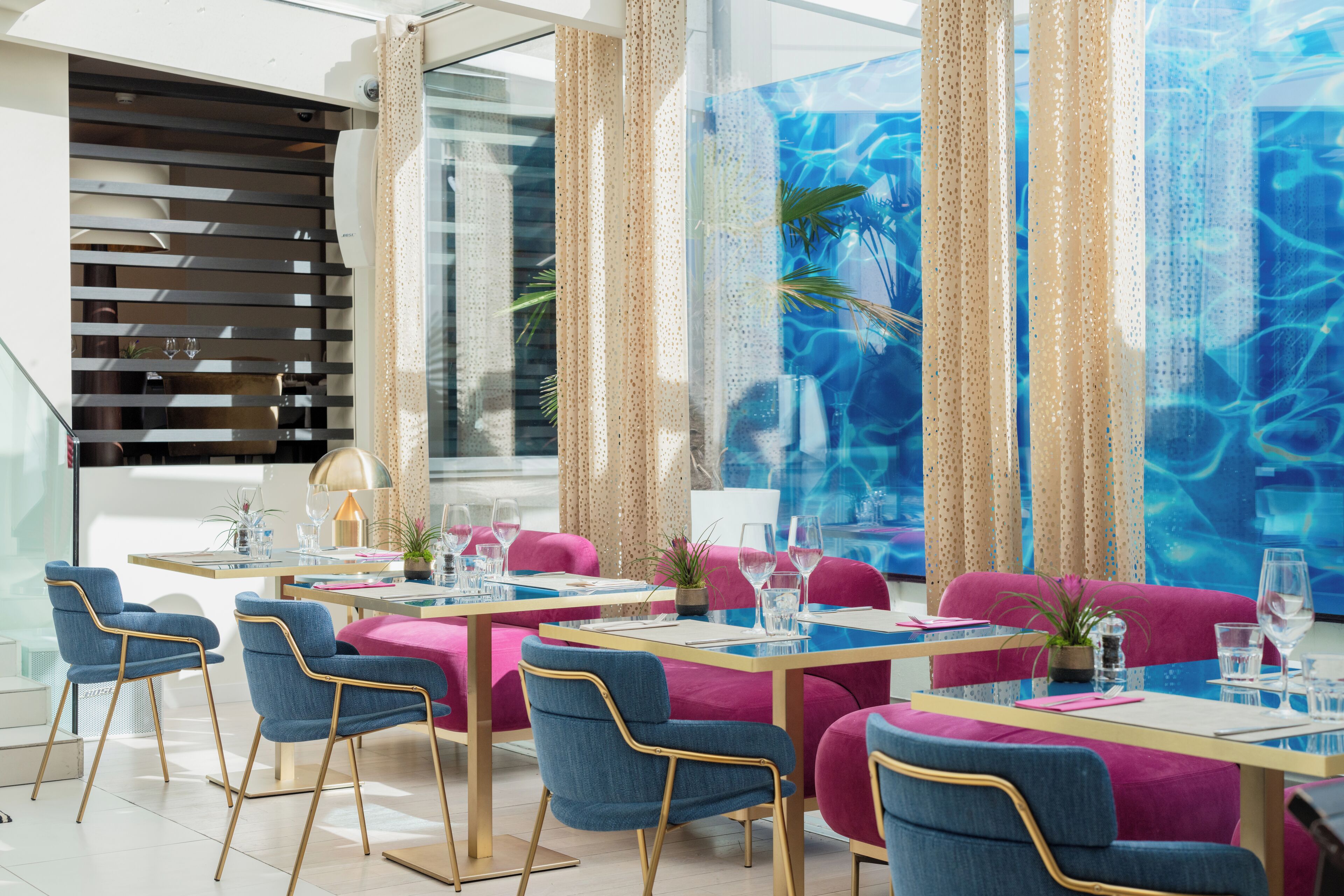 A modern dining area with blue and pink chairs, wooden tables, and a large window with curtains.