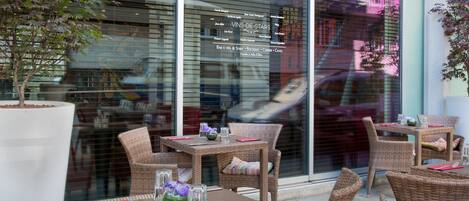 An outdoor seating area with wicker furniture and glassware, a potted plant, and a menu board advertising 'VINS-DE-STARS'.