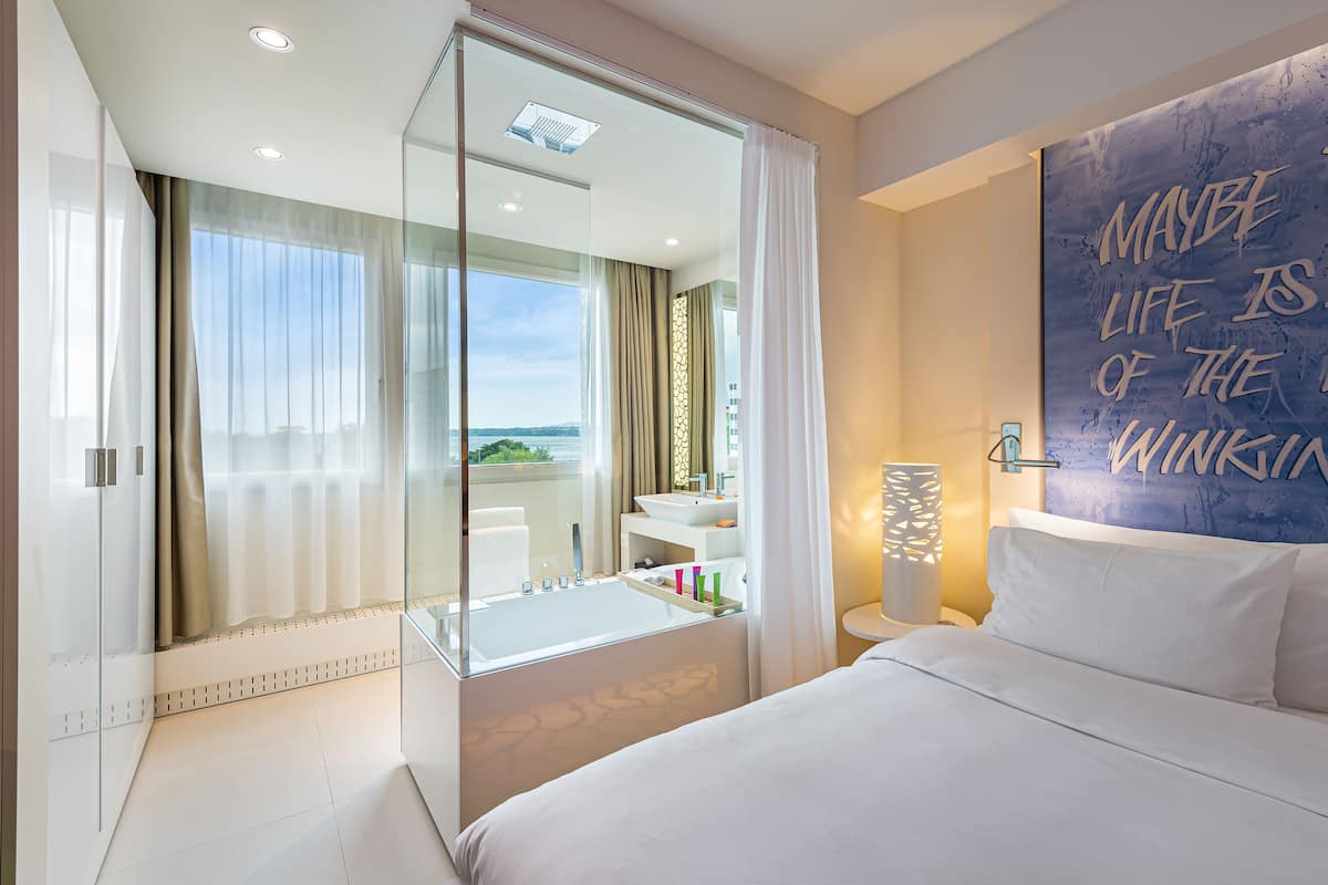 A modern hotel room with a large bed, a glass shower enclosure, a bathroom with a sink and mirror, and a view of the city.