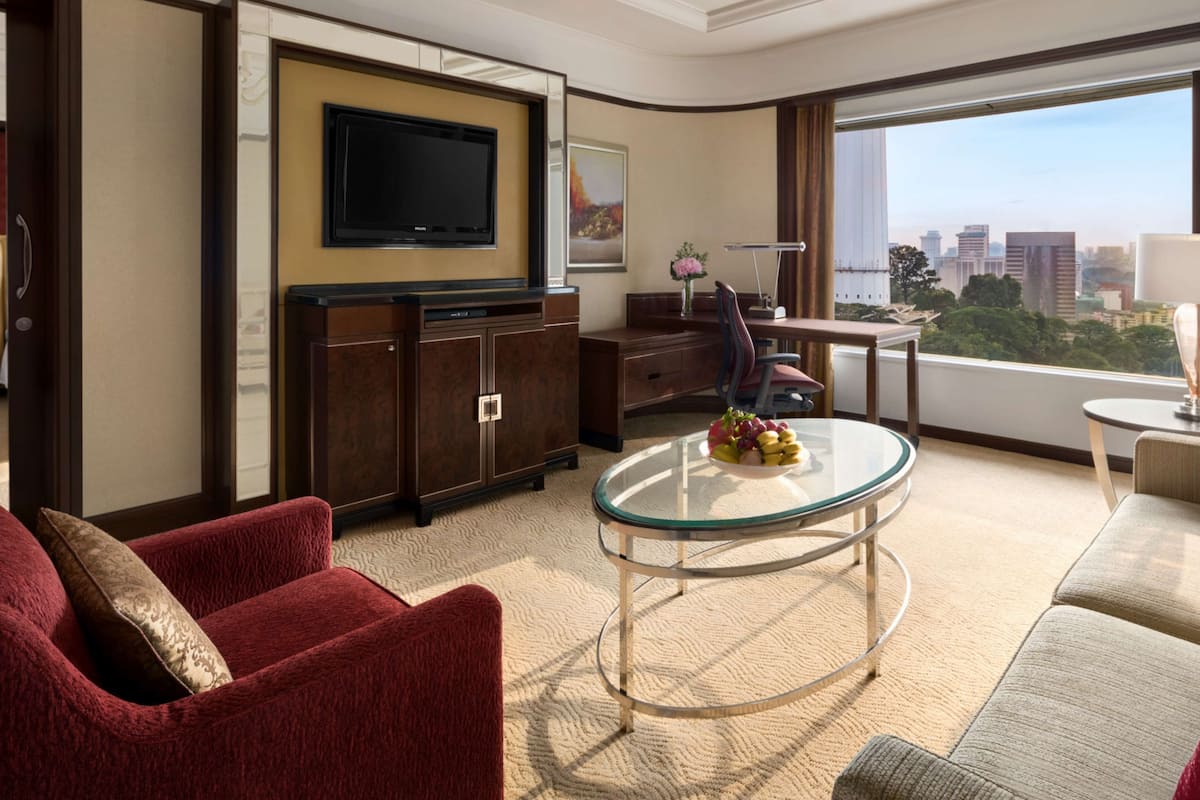 Habitación de hotel con televisor de pantalla plana, comedor con mesa y sillas, sofá, sillón rojo y ventana grande con vista a la ciudad.