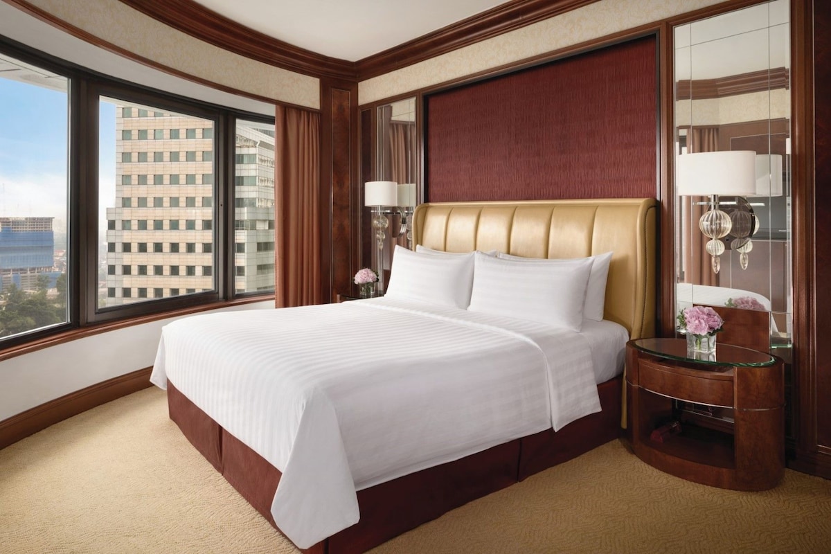 A hotel room with a large bed, a wooden headboard, a nightstand with a lamp, a mirror, and a view of the cityscape.
