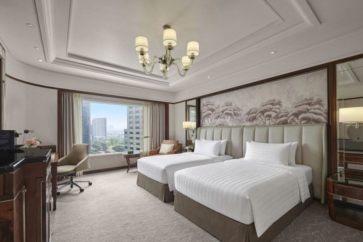A hotel room with two beds, a desk, a chair, a large window, and a chandelier.
