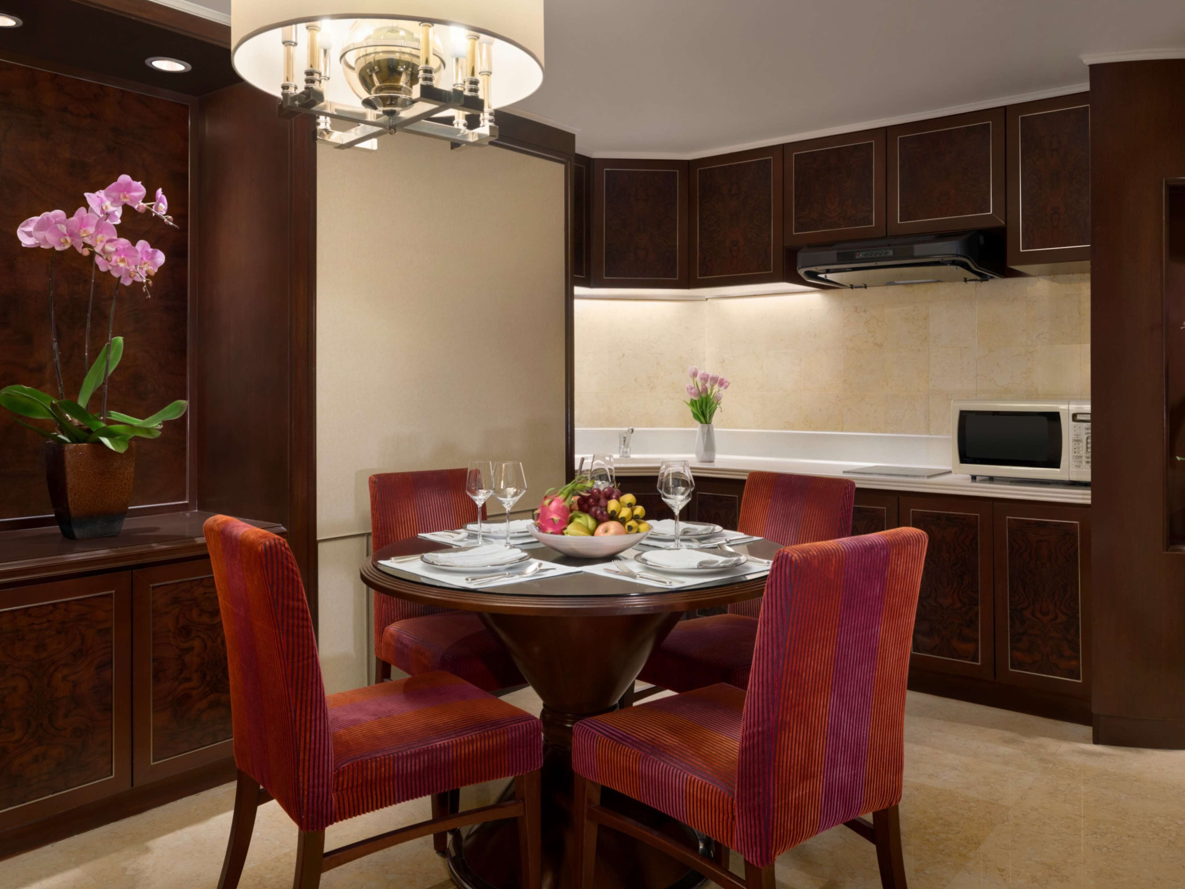 A dining area with a round table, four chairs, a chandelier, a microwave, and a cabinet with a decorative panel.