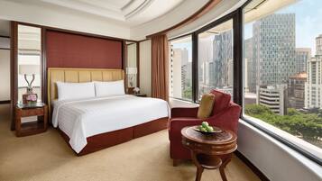 A hotel room with a large bed, a red armchair, a small round table with fruit, and a view of the cityscape through large windows.