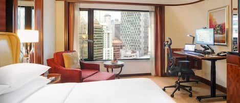 A hotel room with a large bed, a desk with a computer, a chair, and a view of tall buildings.