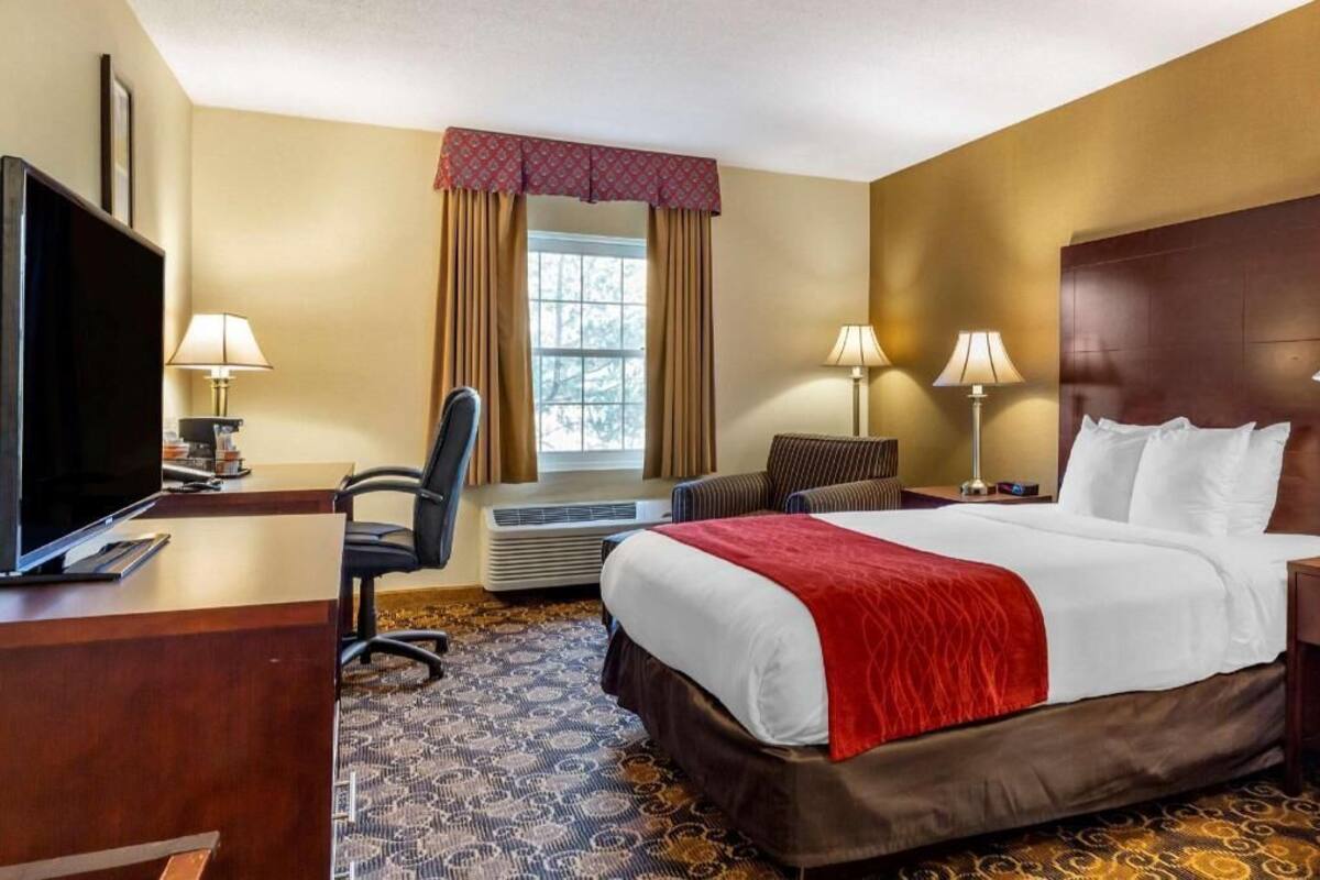 A hotel room with a bed, a desk with a TV, two lamps, a chair, and a window with curtains.