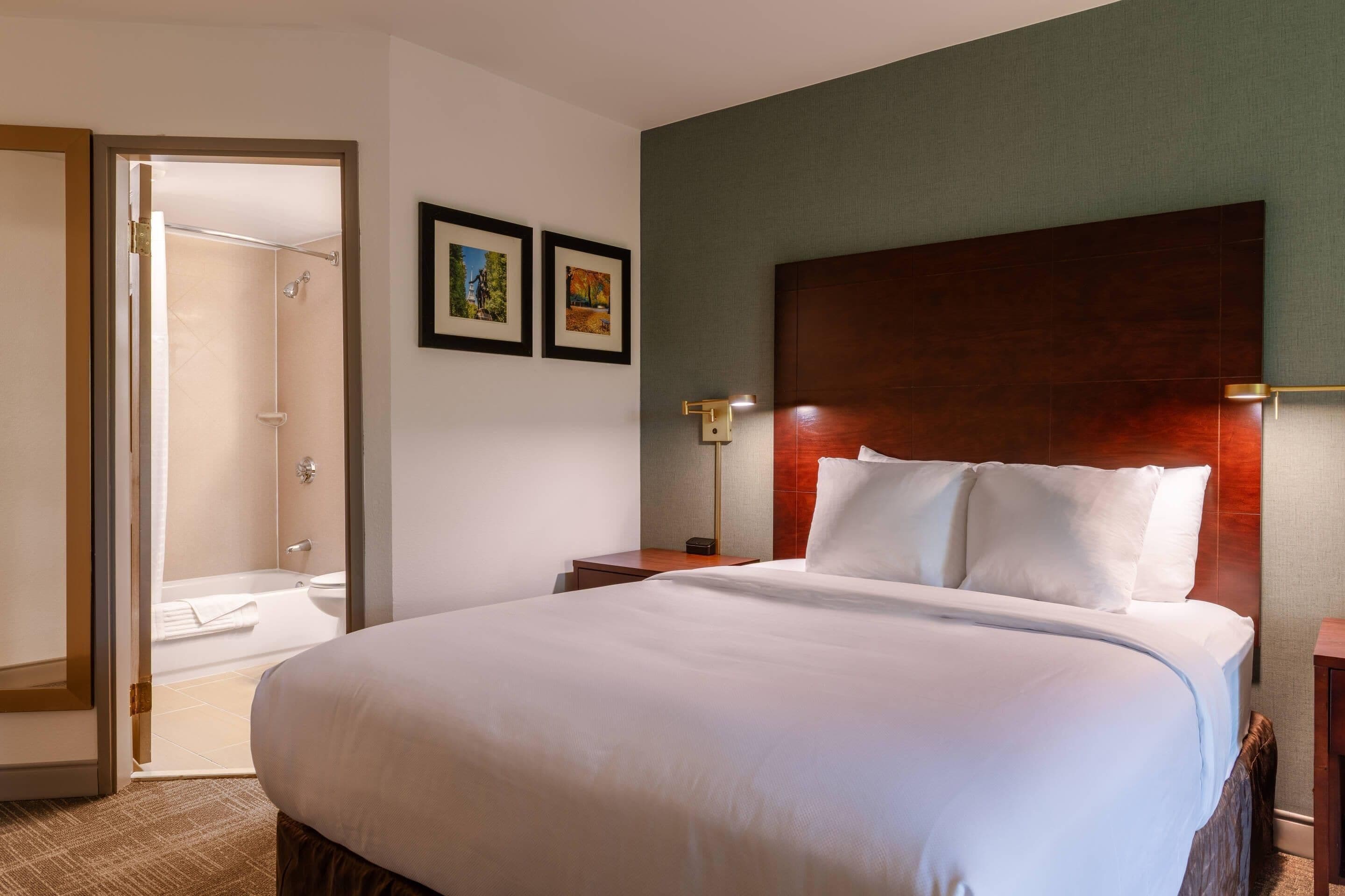 A hotel room with a large bed, a wooden headboard, a nightstand, a wall-mounted lamp, and a framed picture.