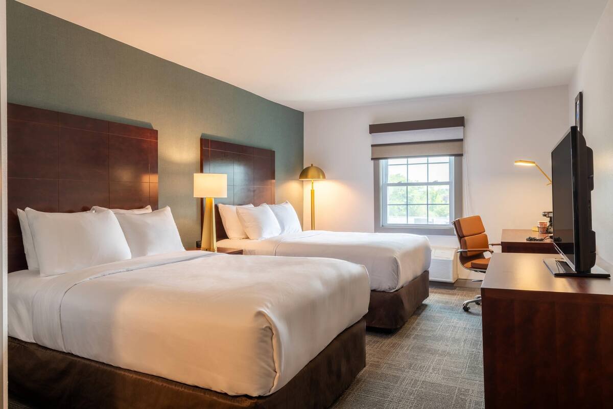 A hotel room with two beds, a desk, a chair, a TV, and a window.