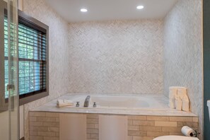 A bathroom with a large bathtub, a window with blinds, and a tiled wall.