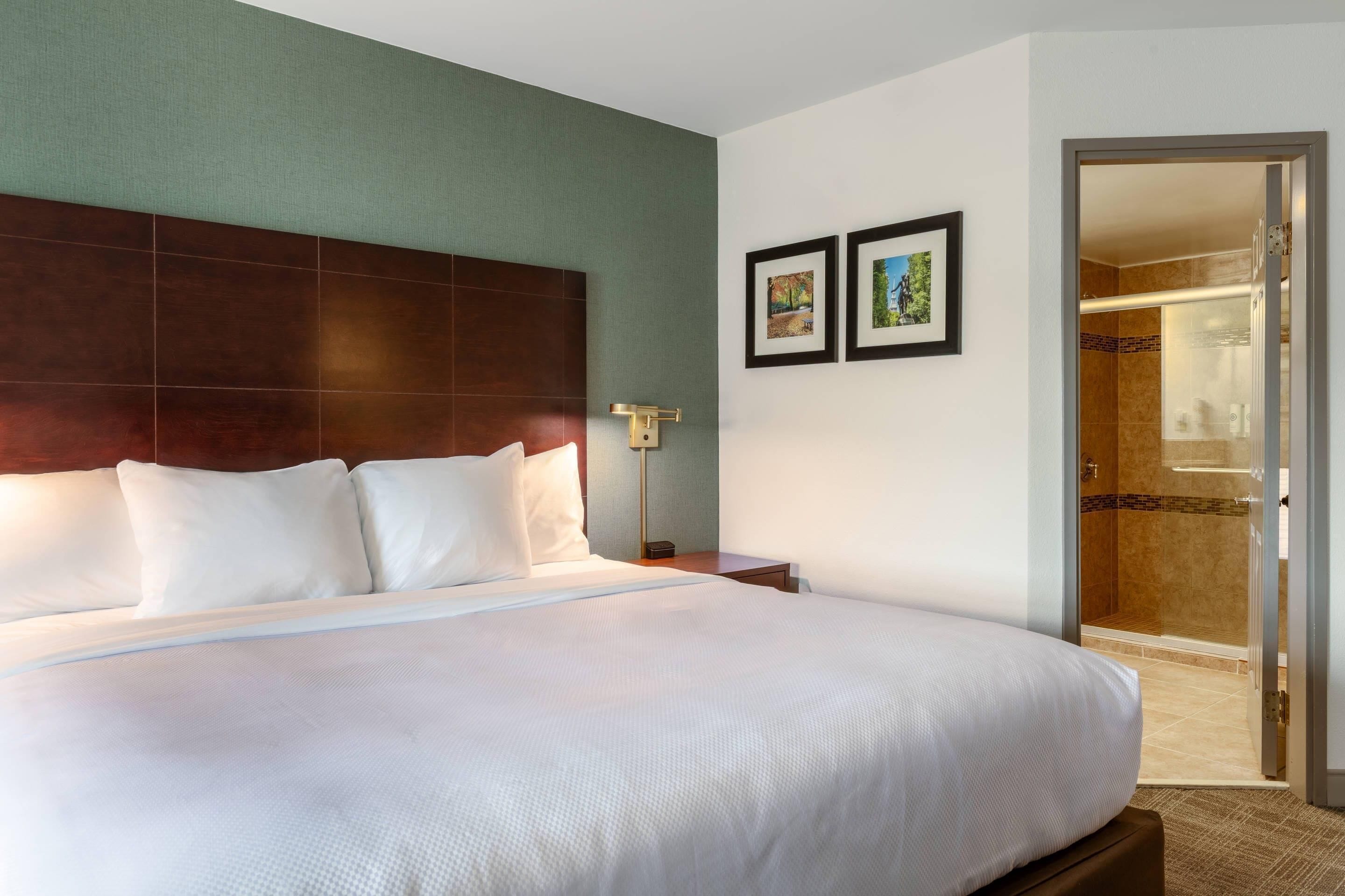 A hotel room with a large bed, a nightstand, a wall-mounted lamp, and a door leading to a shower area.
