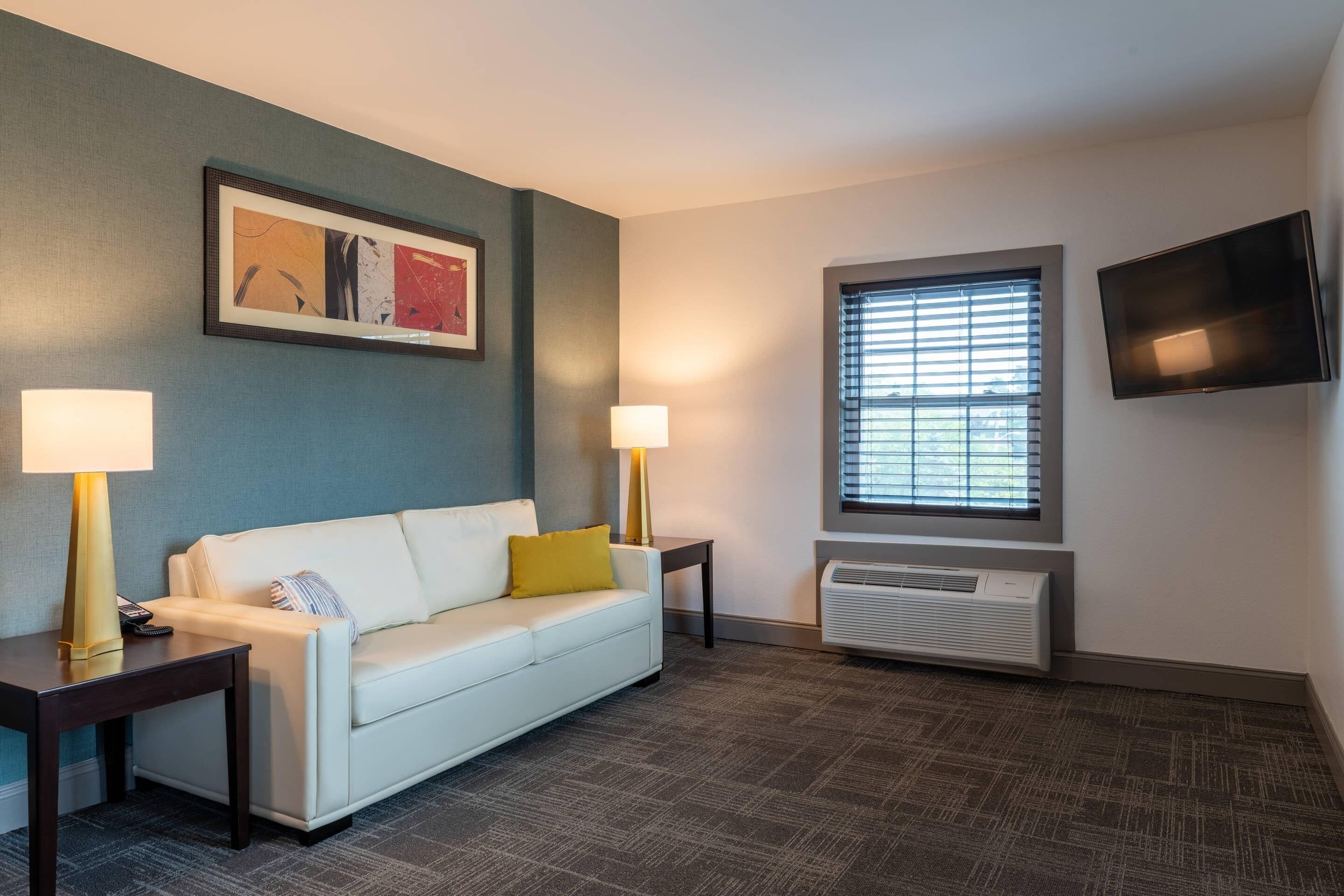 A hotel room with a white sofa, two bedside lamps, a framed abstract painting, a window with blinds, and a wall-mounted TV.