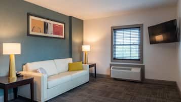 A hotel room with a white sofa, two bedside lamps, a framed abstract painting, a window with blinds, and a wall-mounted TV.