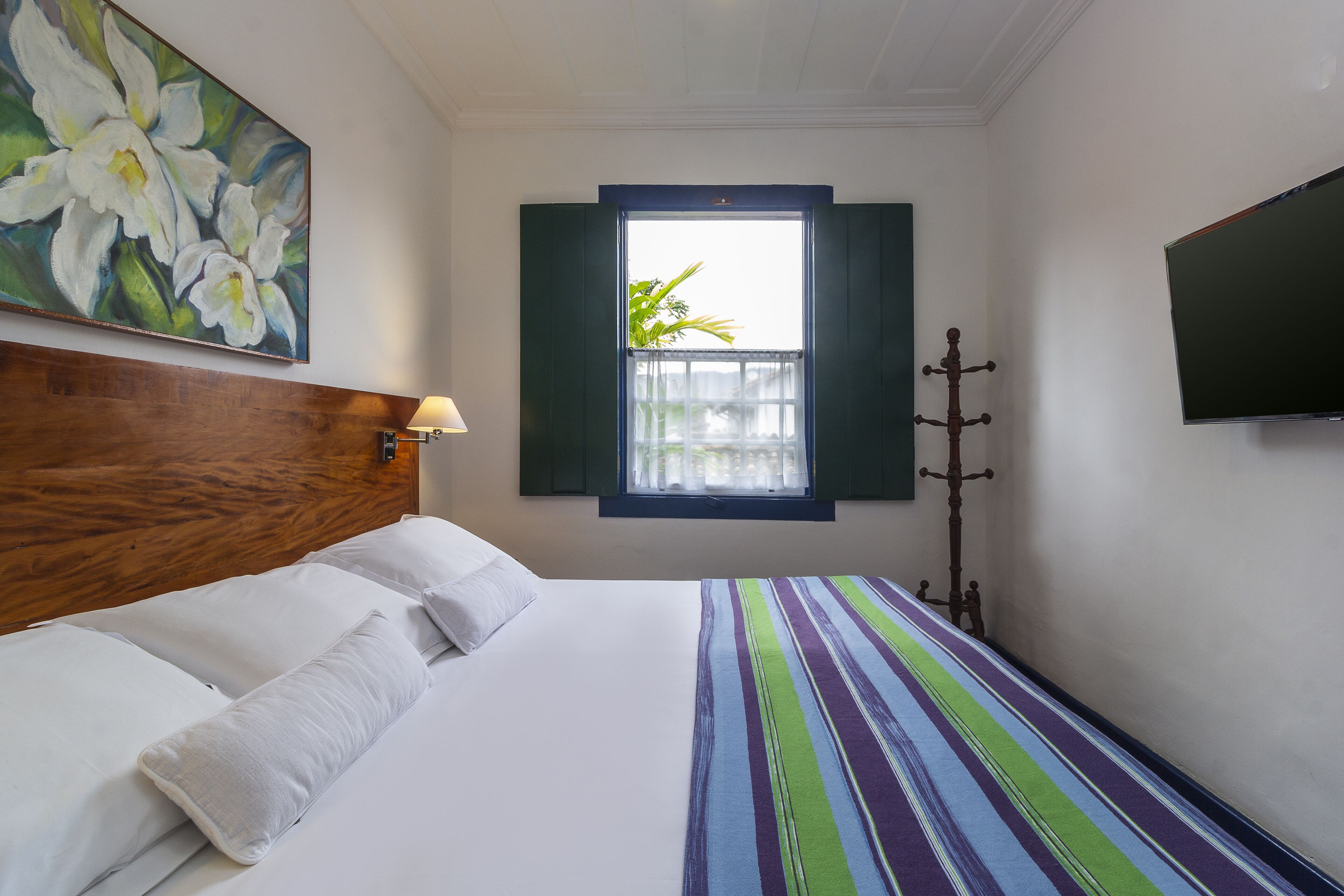 A hotel room with a wooden headboard, a large bed with white linens, a colorful striped bedspread, a window with green shutters, and a flat-screen TV mounted on the wall.