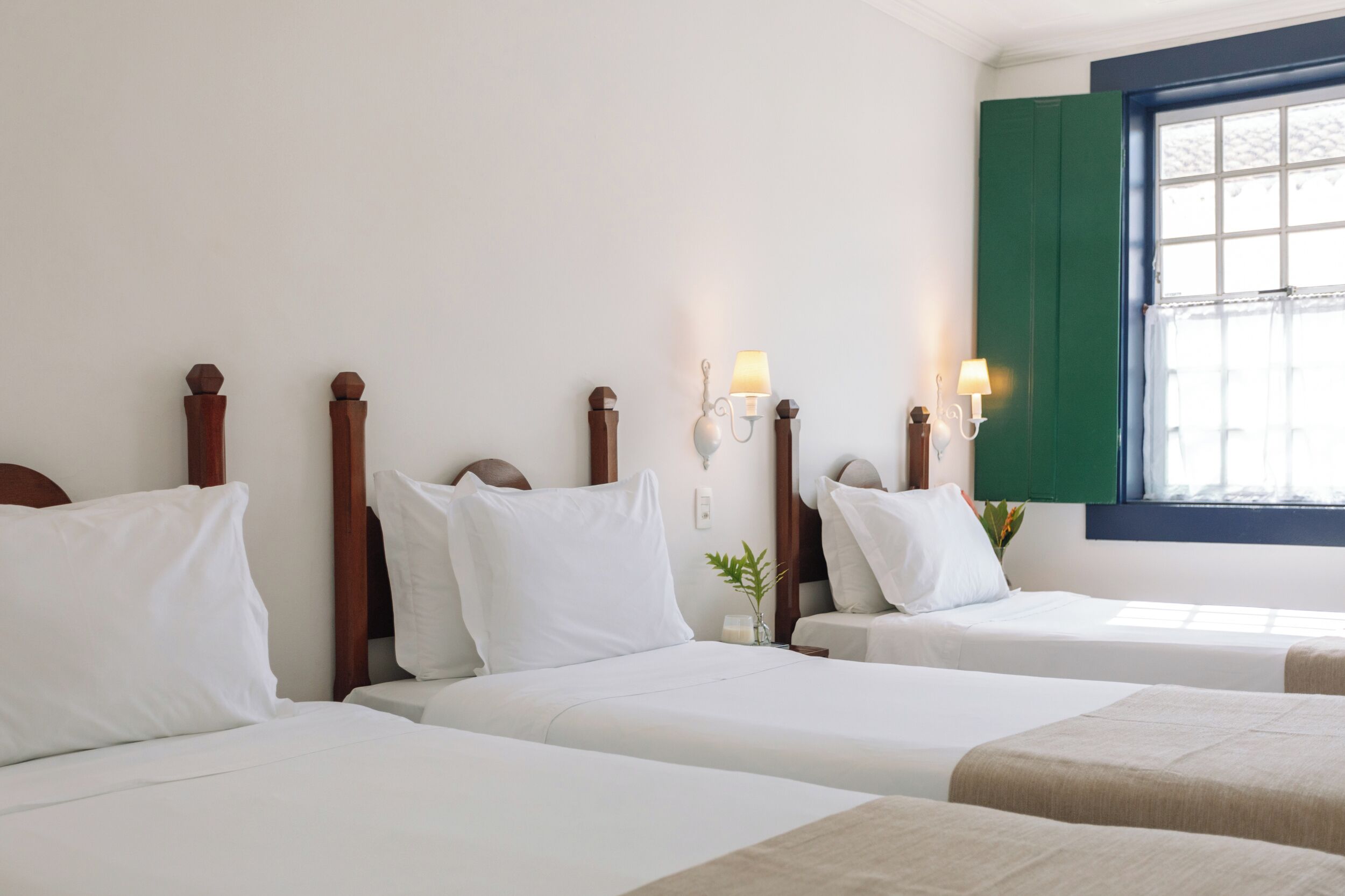 A hotel room with two beds, wooden headboards, white bedding, wall-mounted lamps, and a window with green shutters.