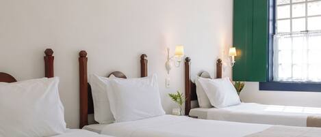 A hotel room with two beds, wooden headboards, white bedding, wall-mounted lamps, and a window with green shutters.