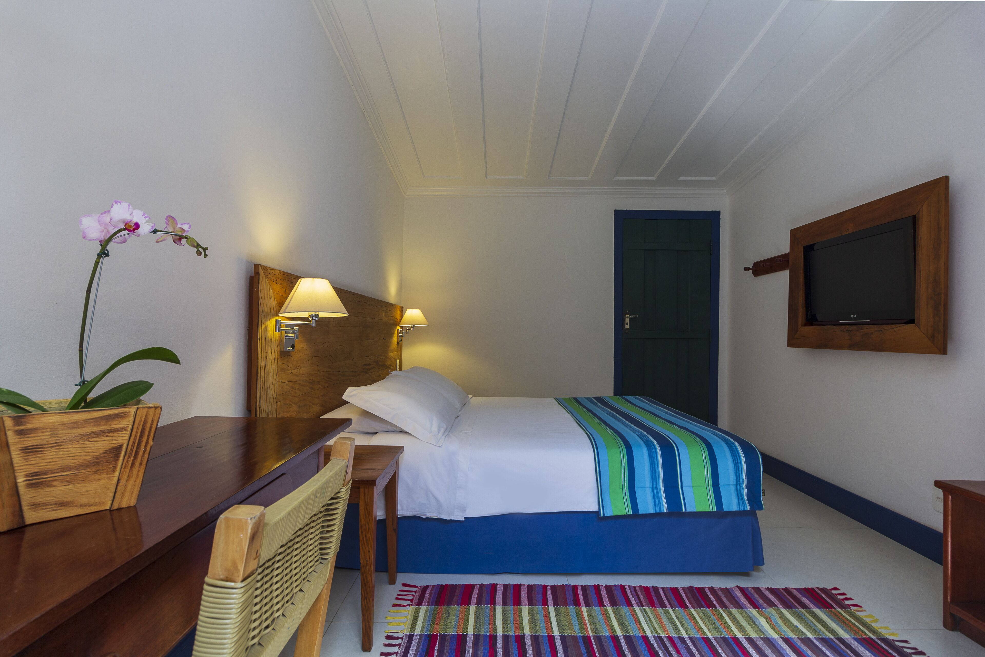Et hotelværelse med en seng, et træsengegærde, sengeborde med lamper, et fjernsyn ophængt på væggen, en farverig stribet måtte og en orkidé i en potte.