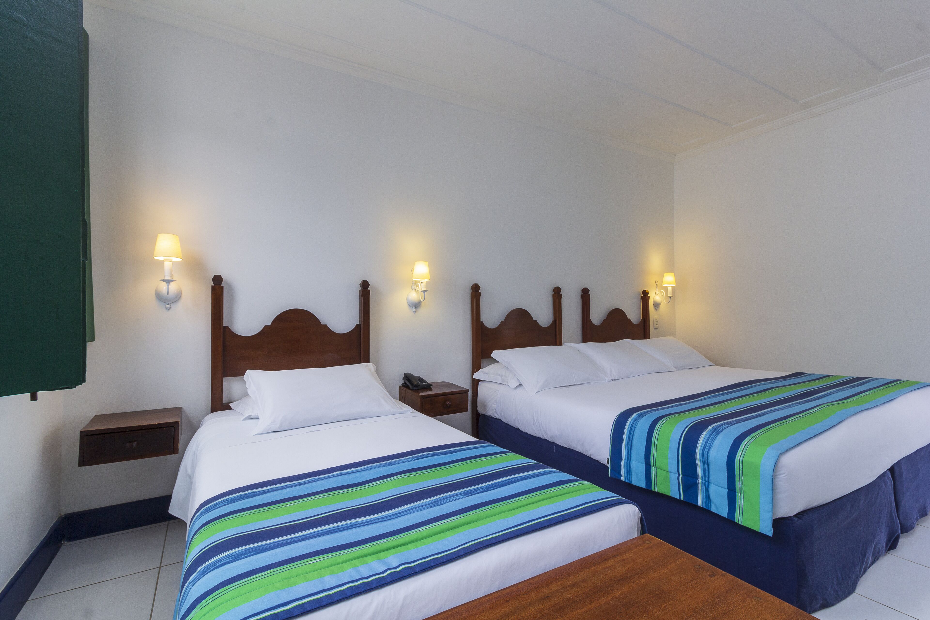 Une chambre double avec deux lits, chacun doté d’une tête de lit en bois, de linge de lit blanc et d’une couette à rayures. On y trouve des tables de chevet avec des lampes et un petit tiroir.