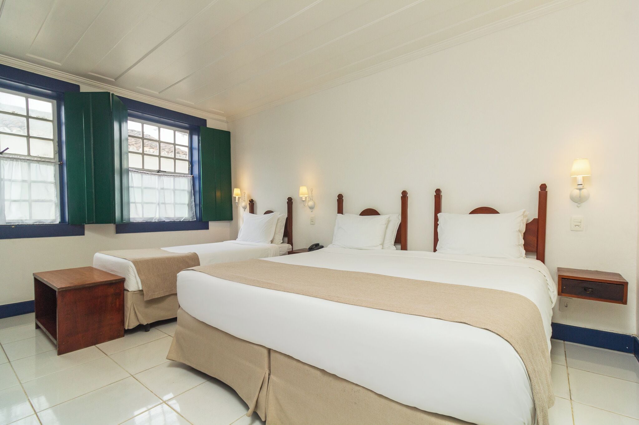 A hotel room with two beds, a small wooden nightstand, and a window with green shutters.