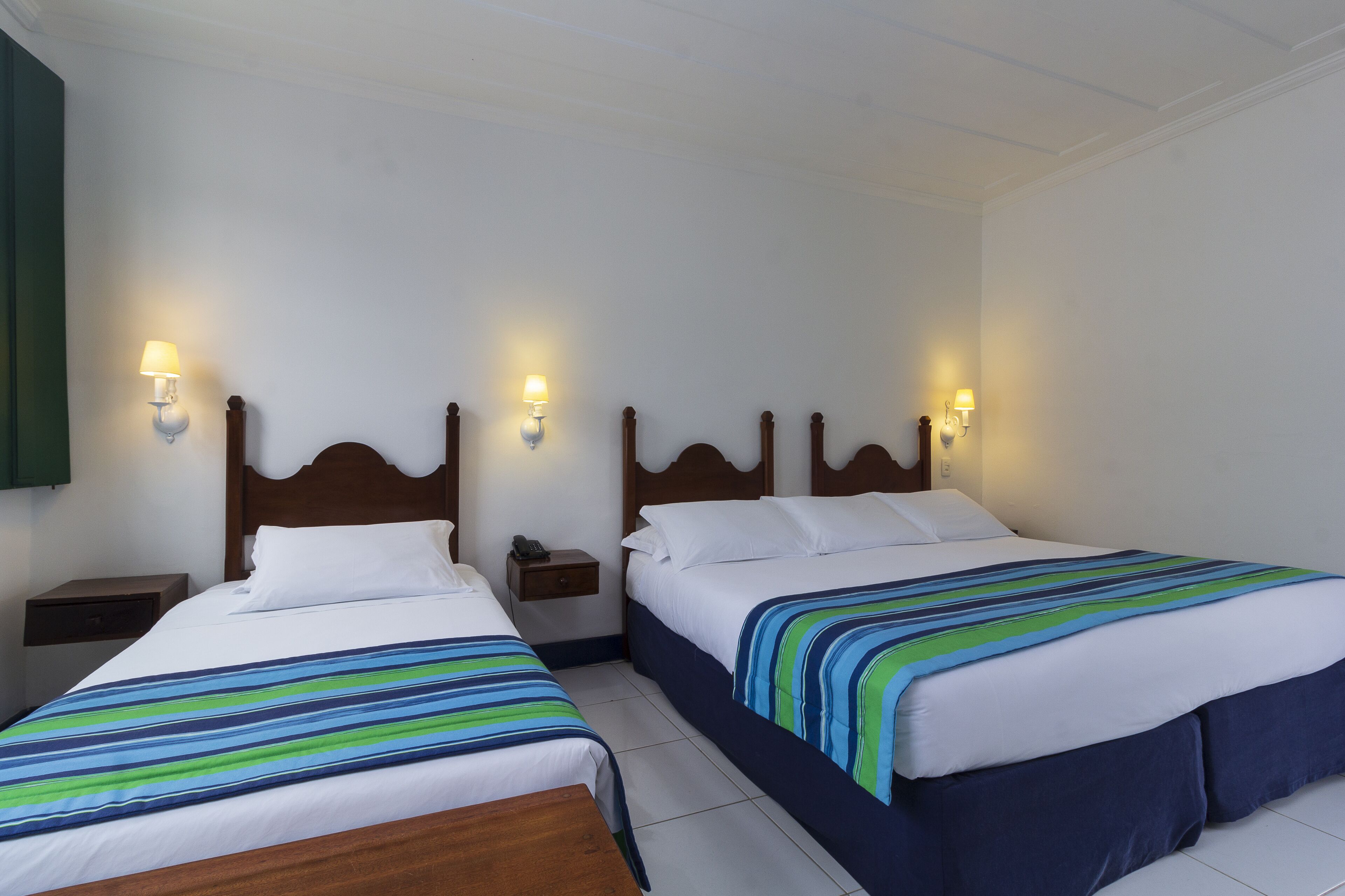 Quarto de hotel com duas camas, cada uma com cabeceira de madeira, roupa de cama branca e cobertores listrados. Há mesas de cabeceira com luminárias e um aplique na parede.