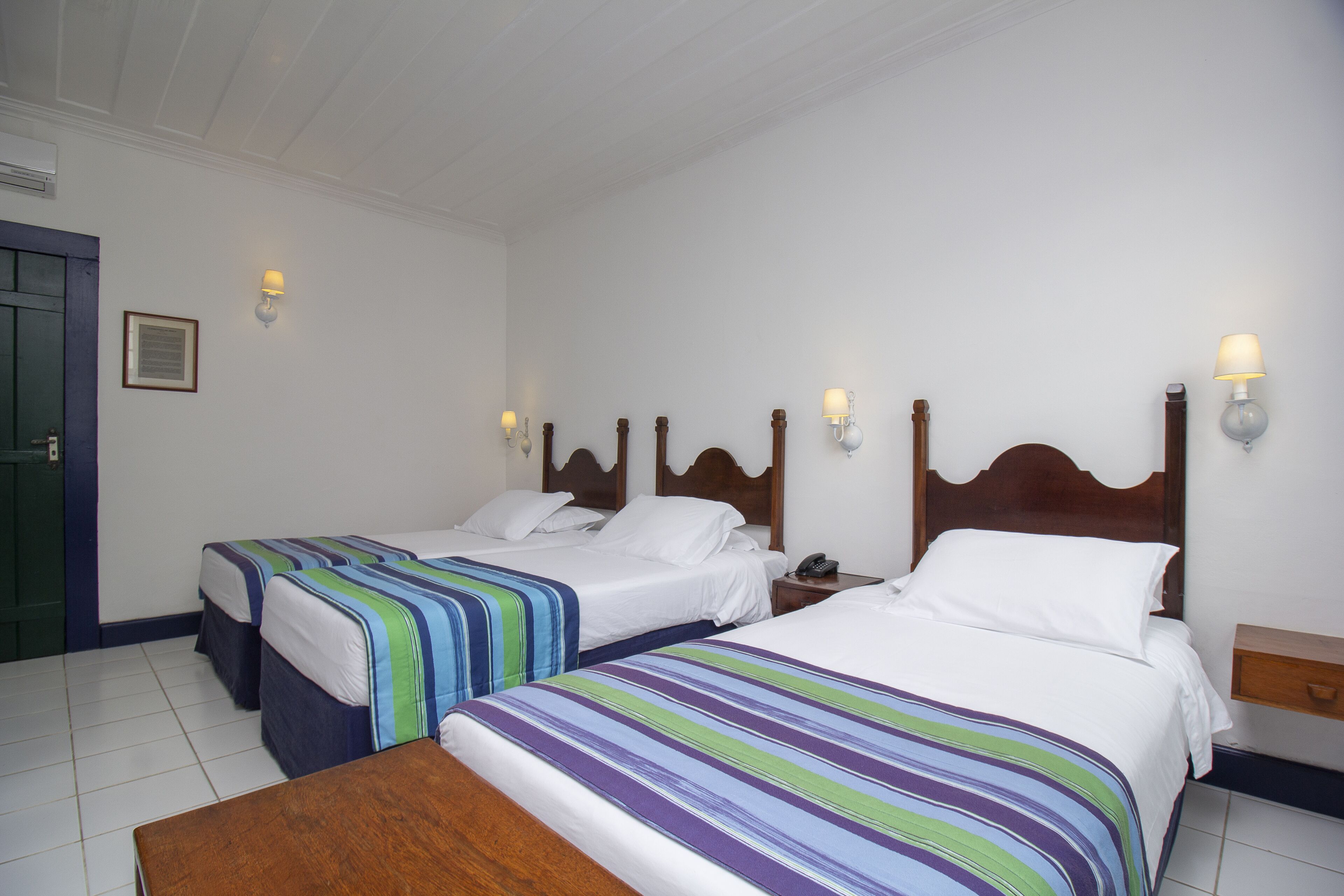 A hotel room with two beds, a wooden headboard, a bedside table with a lamp, and a wooden bench.