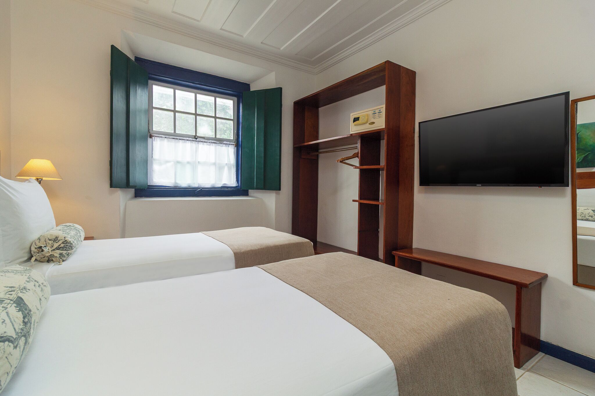 A hotel room with two beds, a window with green shutters, a TV mounted on the wall, and a wooden shelf unit.