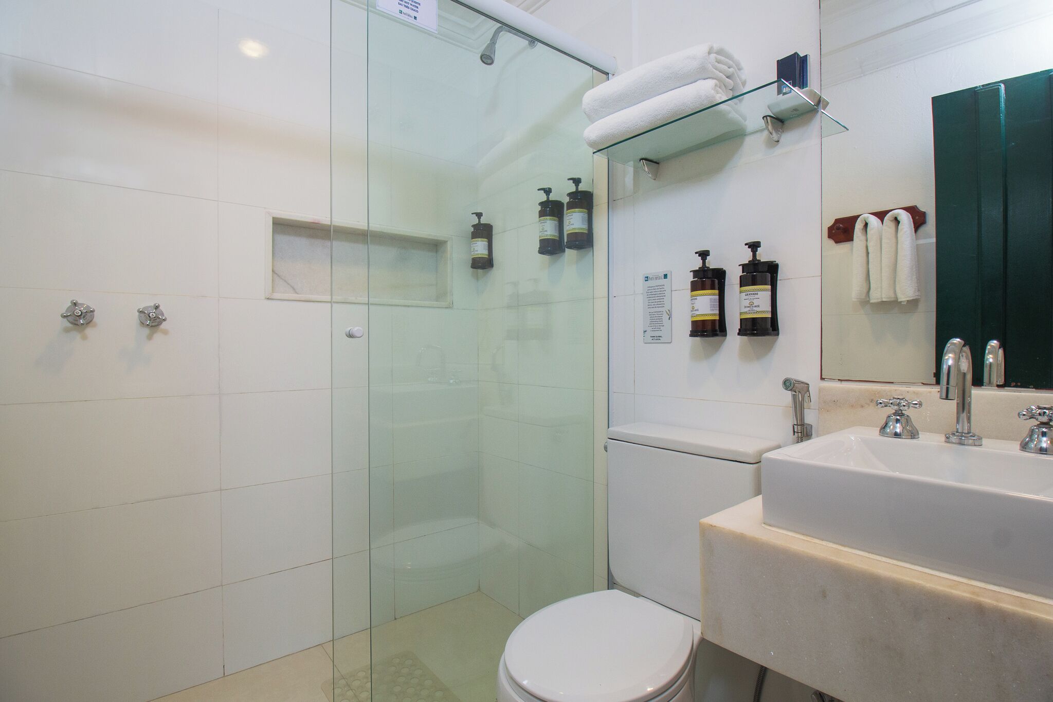 A modern bathroom with a glass-enclosed shower, a white toilet, a sink with two faucets, and toiletries on a shelf.