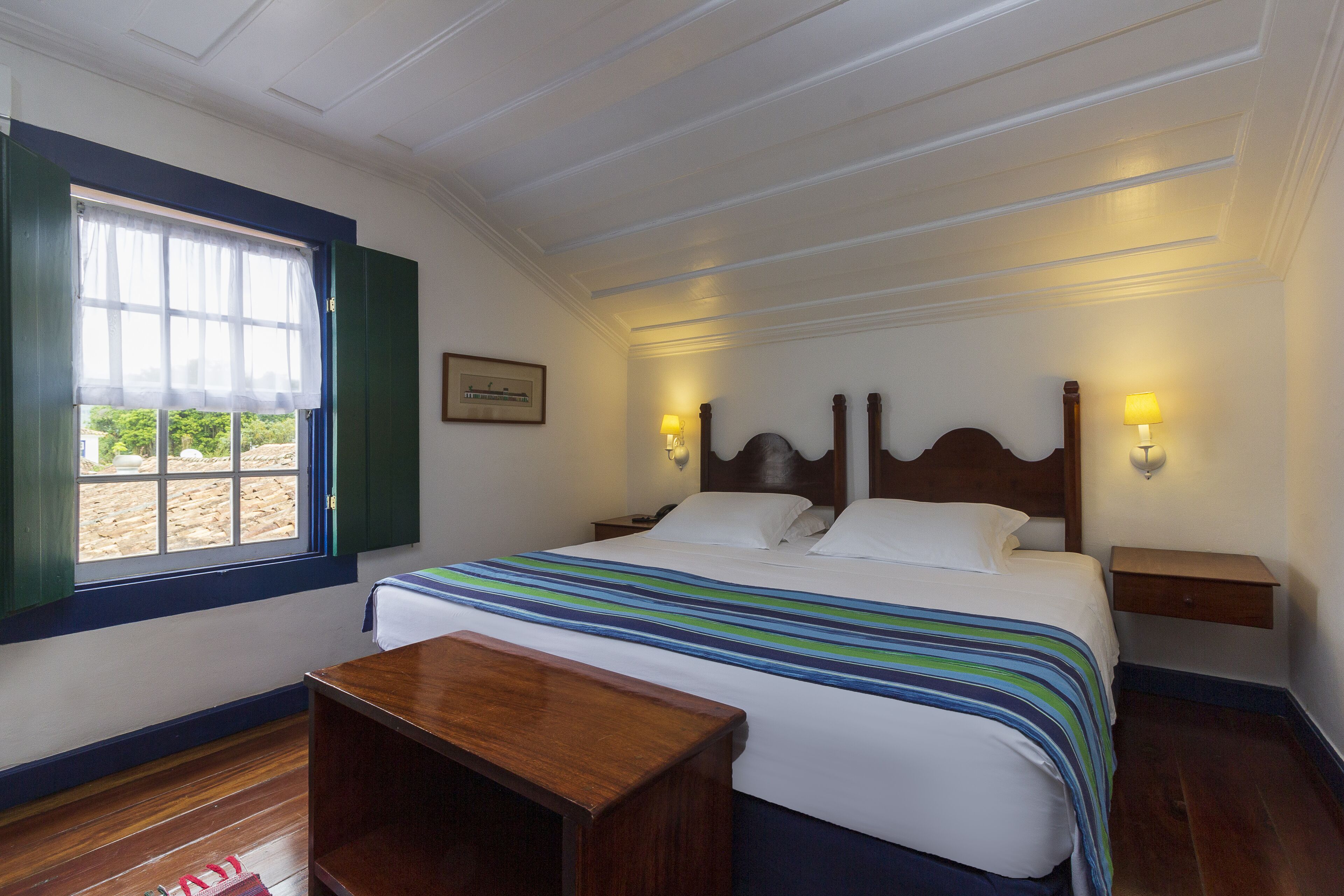 A neatly arranged bedroom with a large bed, bedside tables, a small wooden bench, and a window with a view.