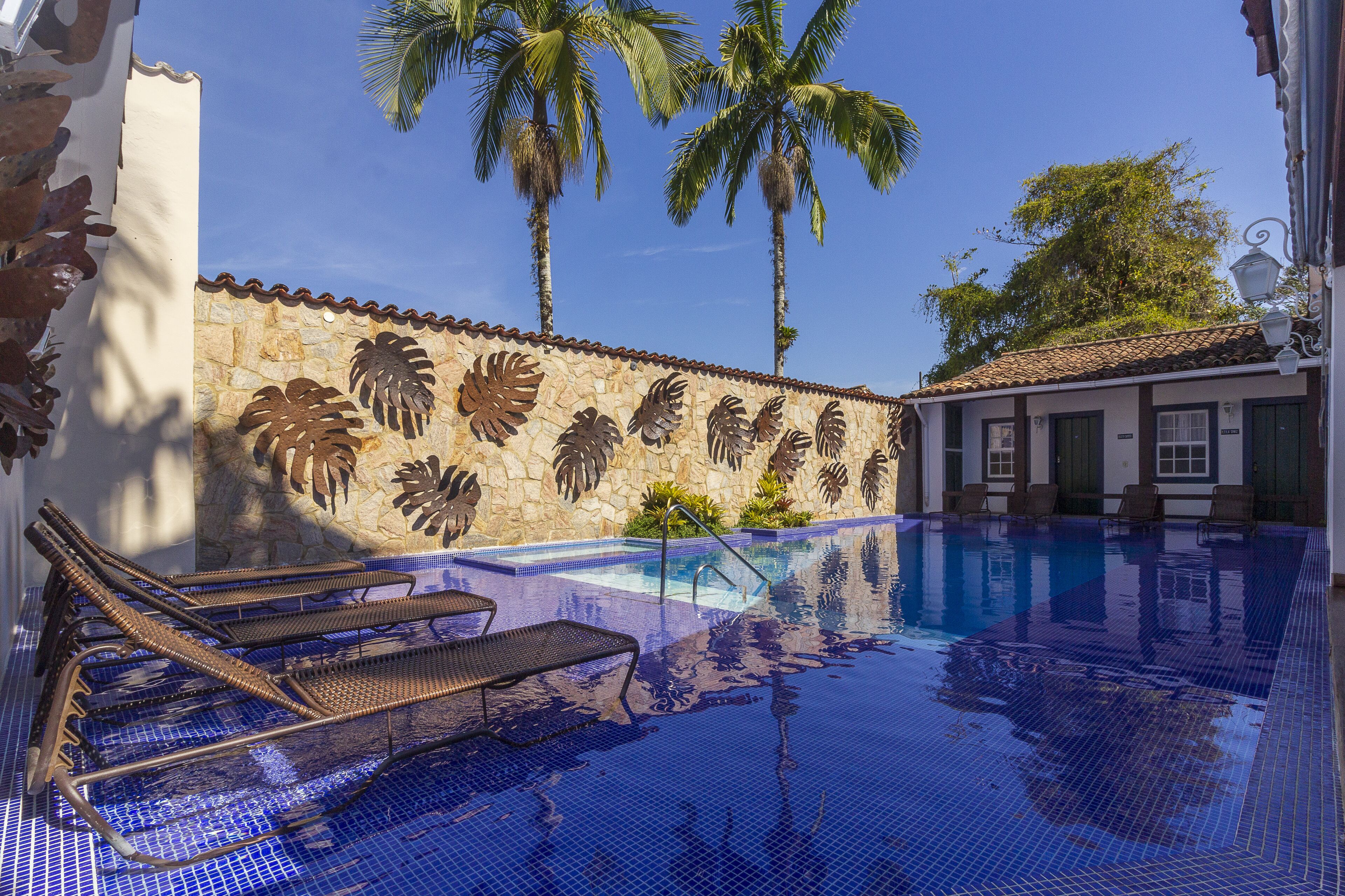 A swimming pool with lounge chairs, palm trees, and a stone wall with leaf patterns.