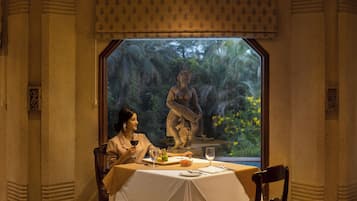 Uma sala de jantar com uma mesa posta para dois, uma estátua ao fundo e uma janela com vista para o verde.