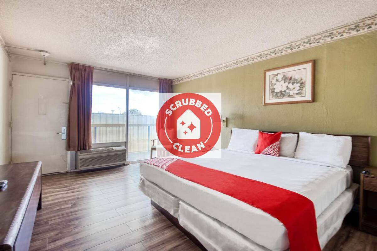 Ein Hotelzimmer mit einem großen Bett, einer Bank, einem Fenster mit Vorhängen und einem gerahmten Bild an der Wand.