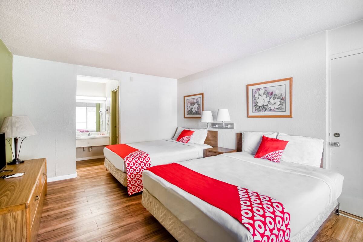 Ein Hotelzimmer mit zwei Betten, jeweils mit einer roten und weißen Tagesdecke, einem Holztisch mit einer Lampe und einem Fenster mit Blick nach draußen.