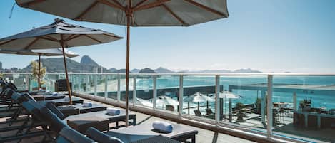 A sunlit outdoor lounge area with lounge chairs, umbrellas, and a view of the ocean.