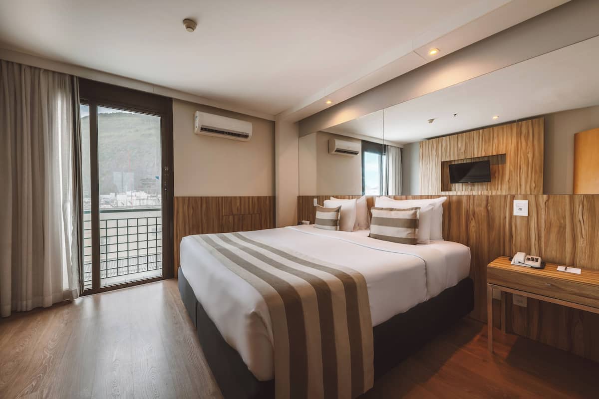 Une chambre d’hôtel avec un grand lit, une table de chevet en bois, une télévision et un balcon avec vue.