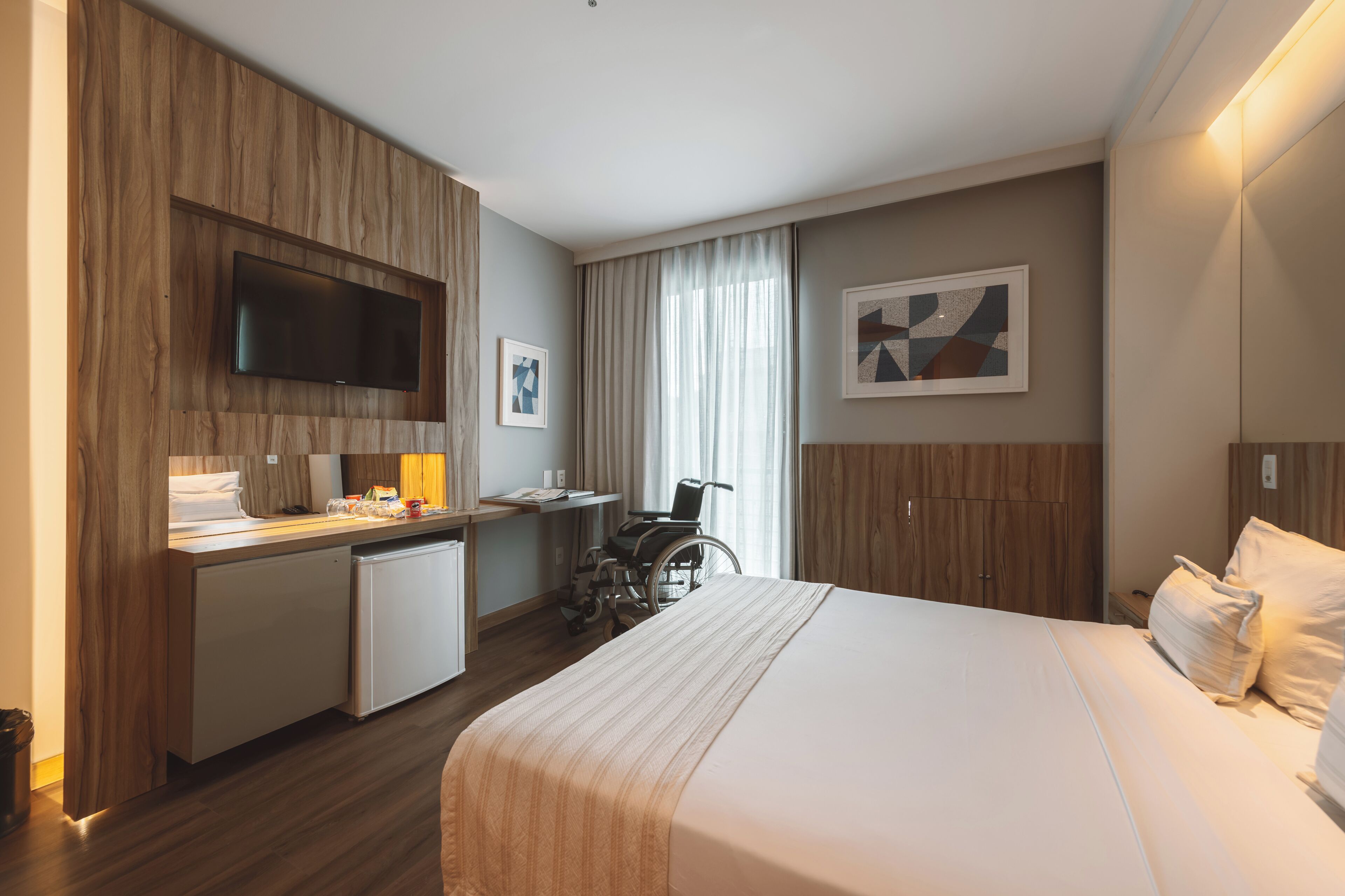 A hotel room with a bed, desk, chair, television, and artwork on the walls.