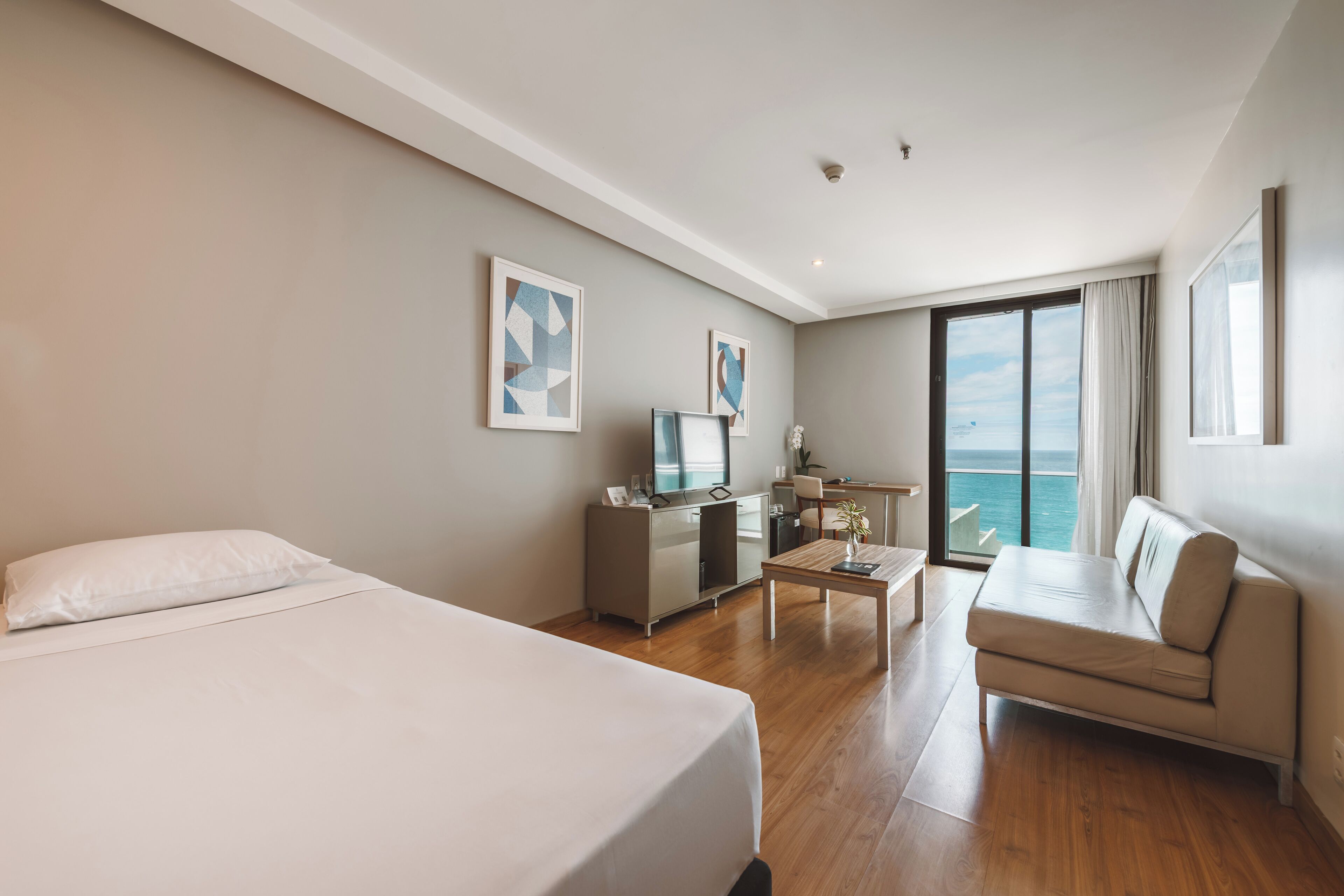 Quarto de hotel com cama, escrivaninha, sofá e uma janela grande com vista.