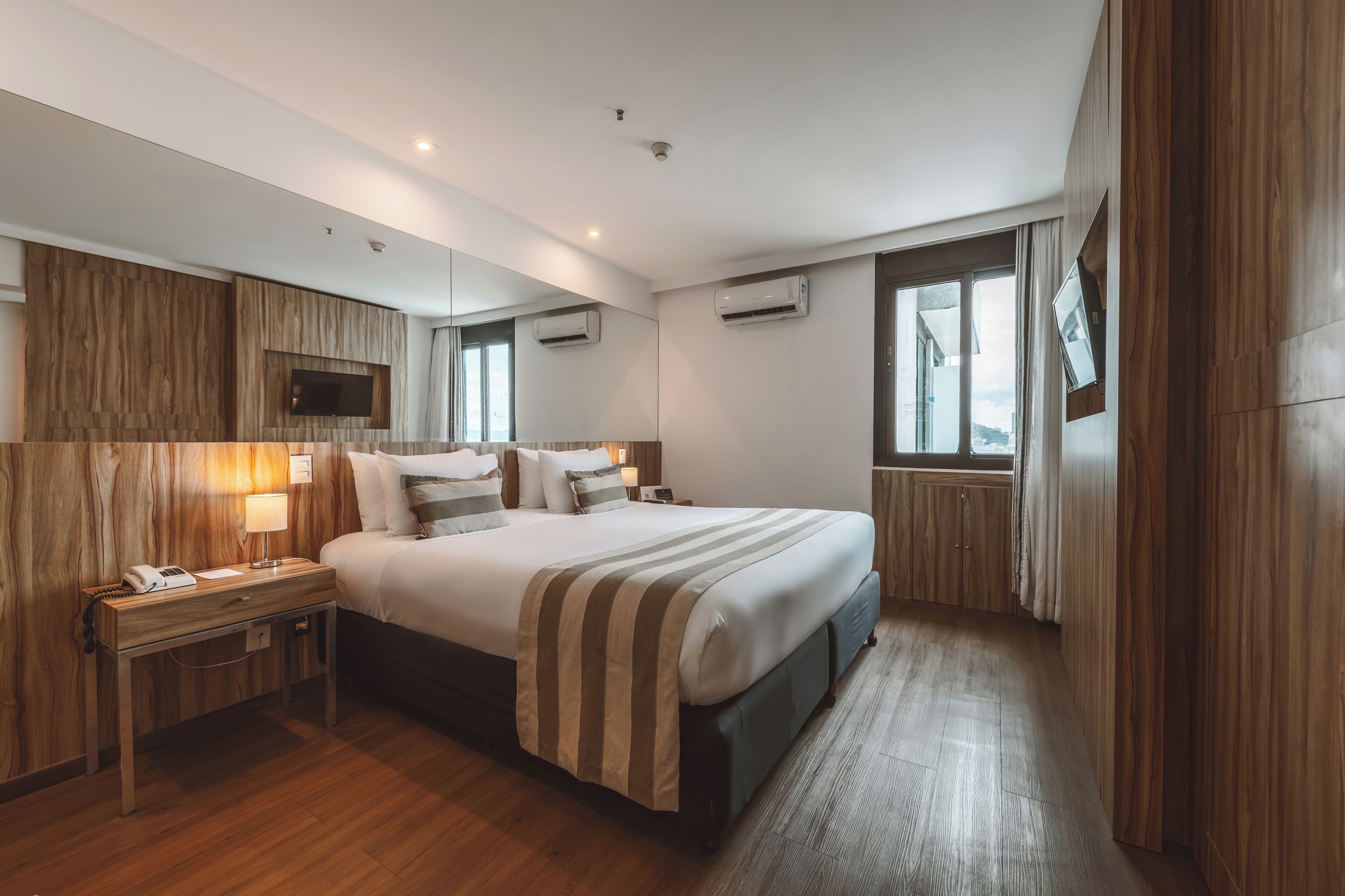 A hotel room with a large bed, a bedside table with a lamp, a TV mounted on the wall, and an air conditioning unit.