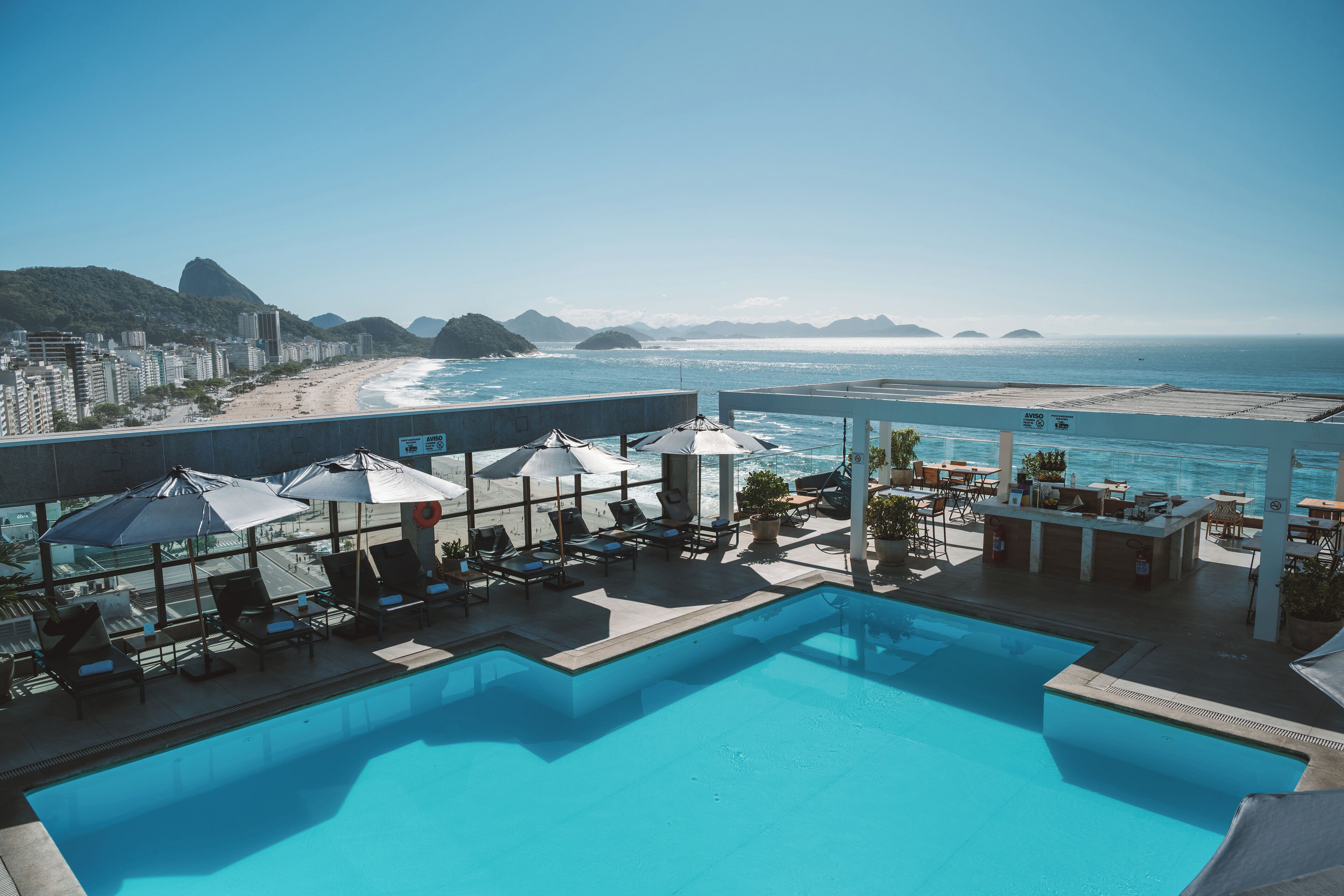 A rooftop pool with lounge chairs and umbrellas, overlooking a beach and cityscape.