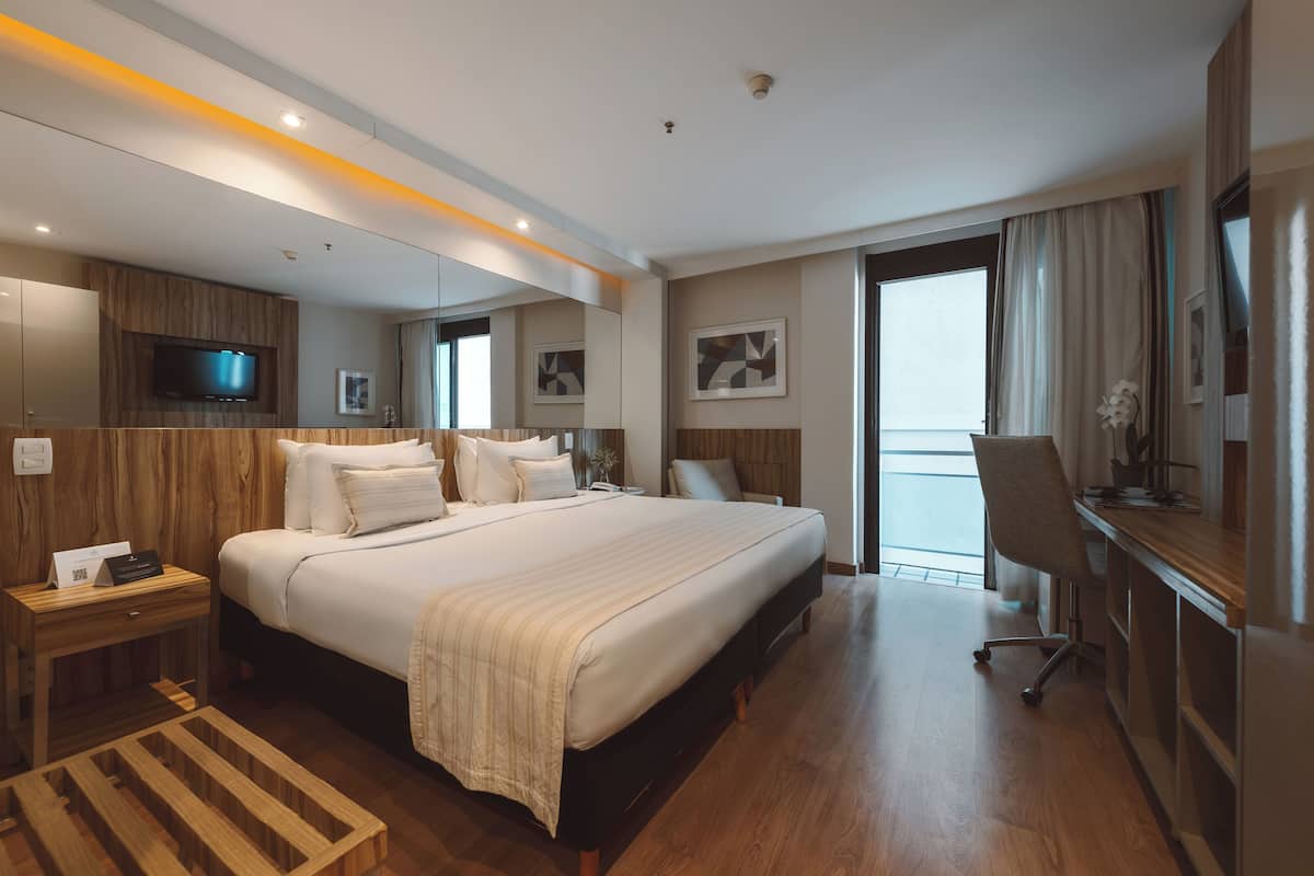 Une chambre d’hôtel avec un grand lit, un bureau, une chaise, une télévision et un balcon avec des rideaux.