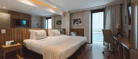 A hotel room with a large bed, a desk, a chair, a TV, and a balcony with curtains.
