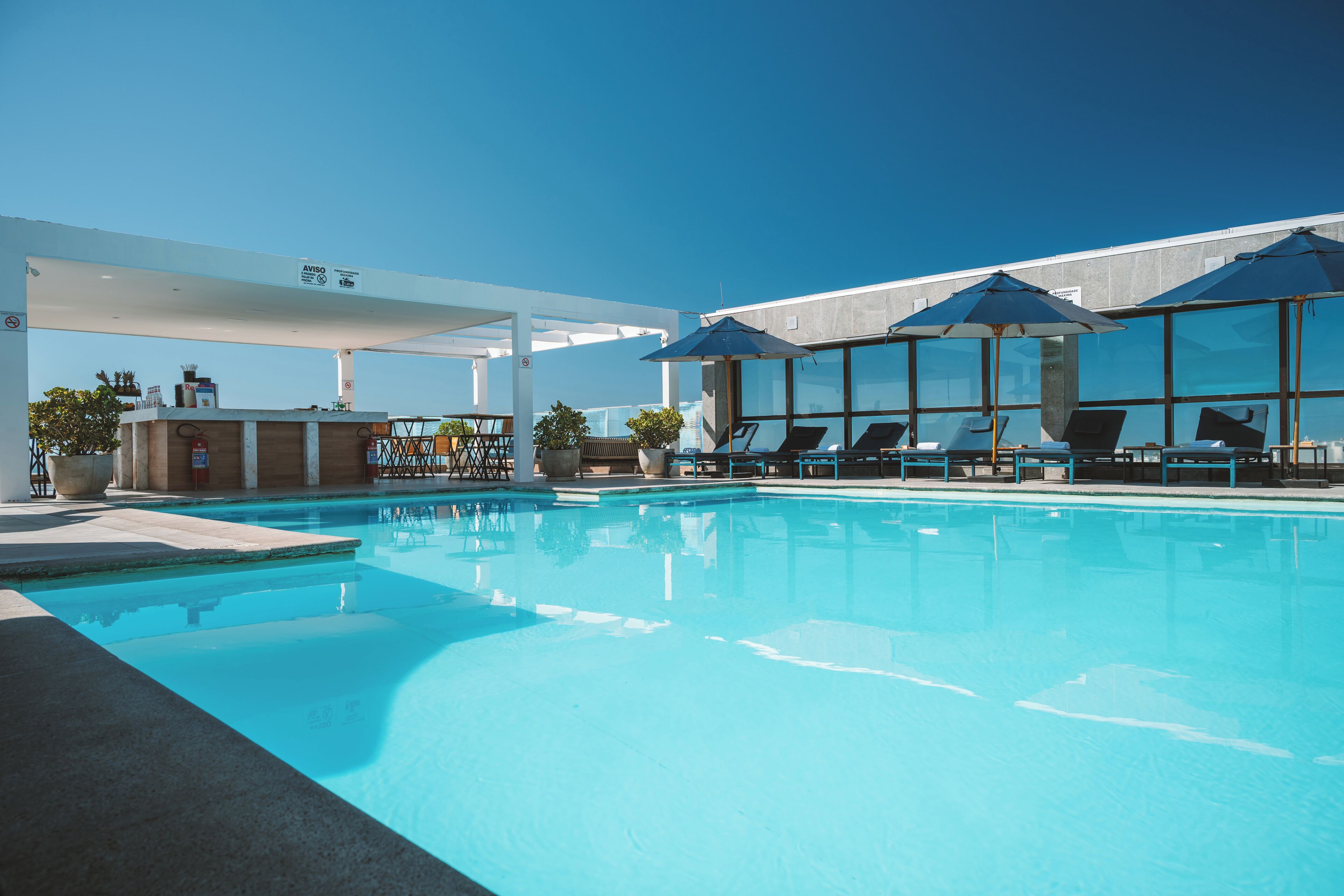 Piscina con sillones de descanso y sombrillas, una zona de bar y un cielo azul despejado.
