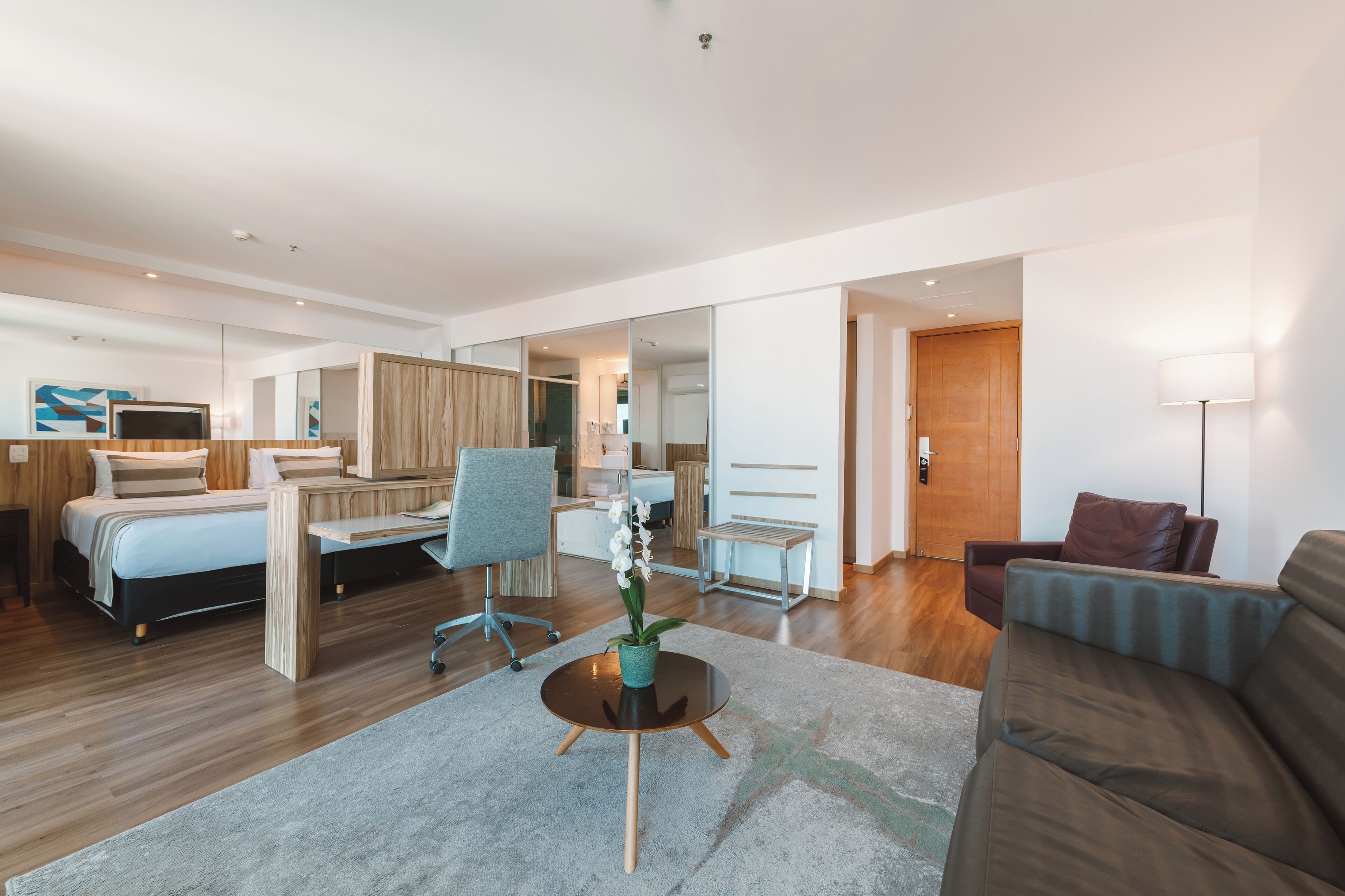 A modern hotel room with a bed, desk, chair, sofa, and a small table with a plant.