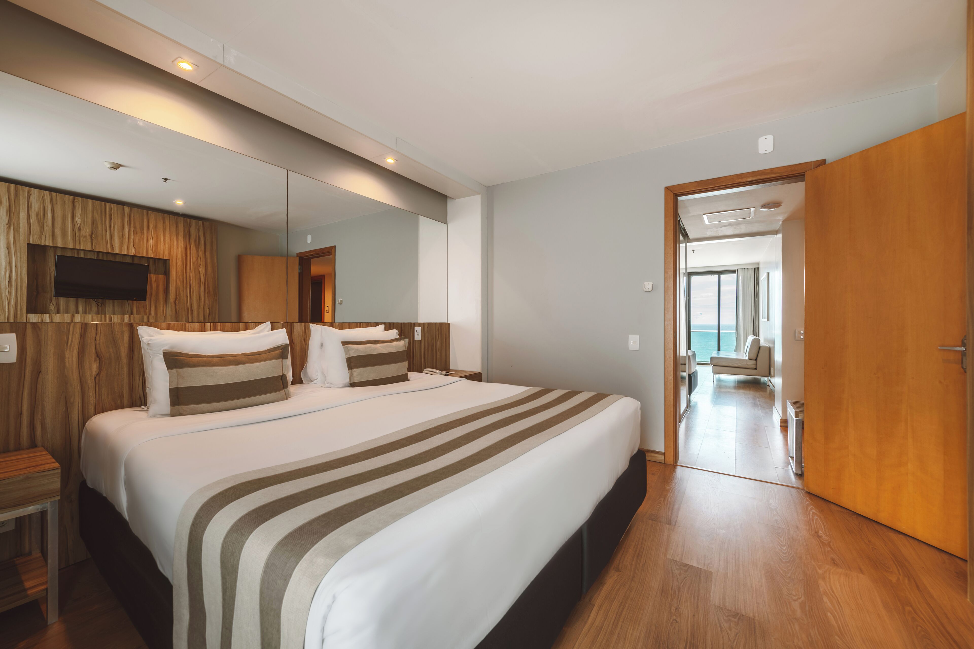 Habitación de hotel con una cama grande, paneles de madera, un televisor de pantalla plana y vista al mar a través de un ventanal.